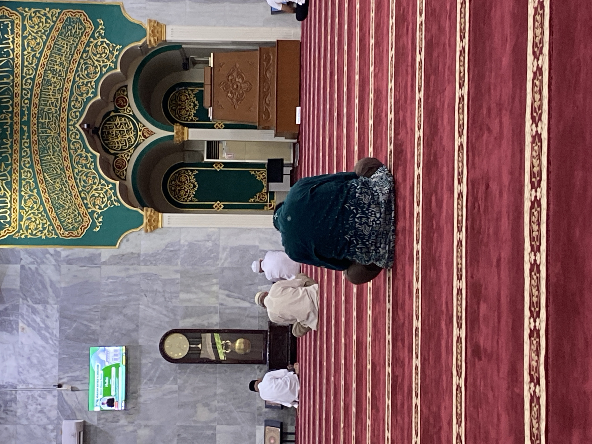 The image depicts the interior of a mosque in Lhokseumawe, Indonesia. In the foreground, a person is seen from the back, seated on a red carpet with striped patterns. Several other individuals, also facing away from the camera, are seated on the carpet in the middle ground, appearing to be engaged in prayer or religious observance. The background features a large, ornately decorated archway with Islamic calligraphy and geometric patterns in green and gold. A wooden pulpit, a grandfather clock, and a television screen displaying an image are also visible against a marble-tiled wall. The overall environment suggests a place of worship, likely during a prayer service.