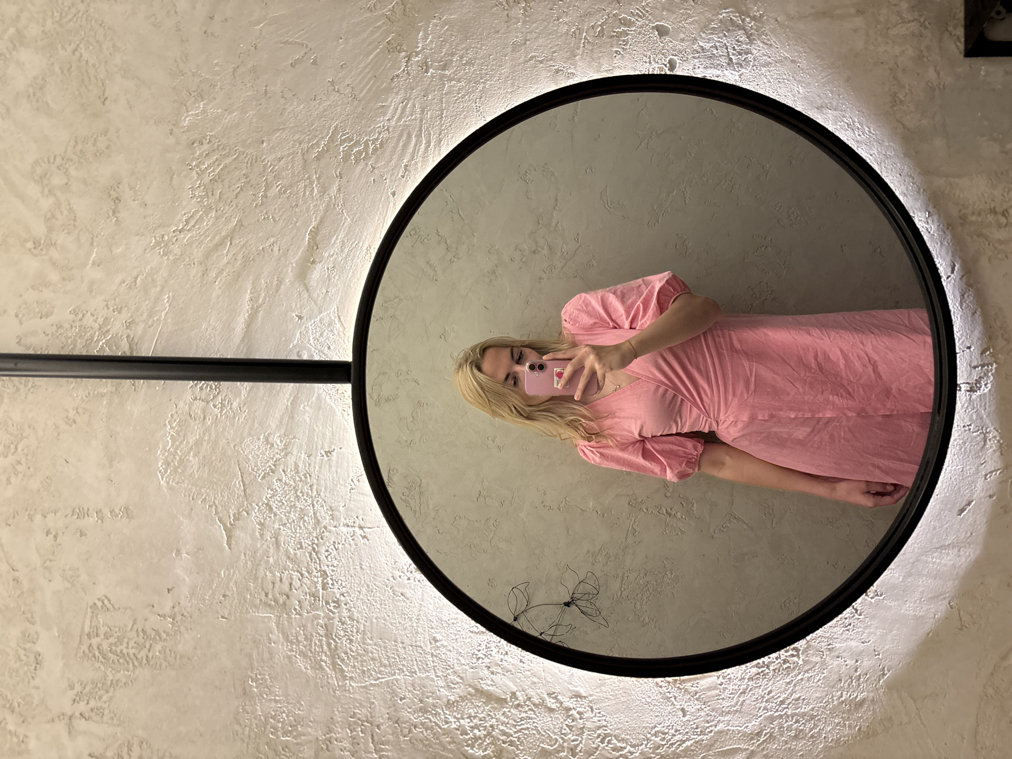 An indoor scene in Krasnoyarsk, Russia, captures a blonde woman reflected in a large, circular, black-framed mirror that features an internal backlight, illuminating the surrounding wall. The woman wears a pink wrap dress with short, puffy sleeves and holds a pink smartphone in her right hand, angled as if taking a selfie, with her eyes directed at the phone screen. A small pink accessory is visible on the back of the phone. The wall, both around the mirror and reflected within it, displays a heavily textured, light beige or off-white plaster finish with irregular raised patterns. To the left side within the mirror's reflection, a minimalist wire art piece depicting two floral outlines is affixed to the wall. A black vertical pole or support structure extends upwards from the top center of the mirror's frame.