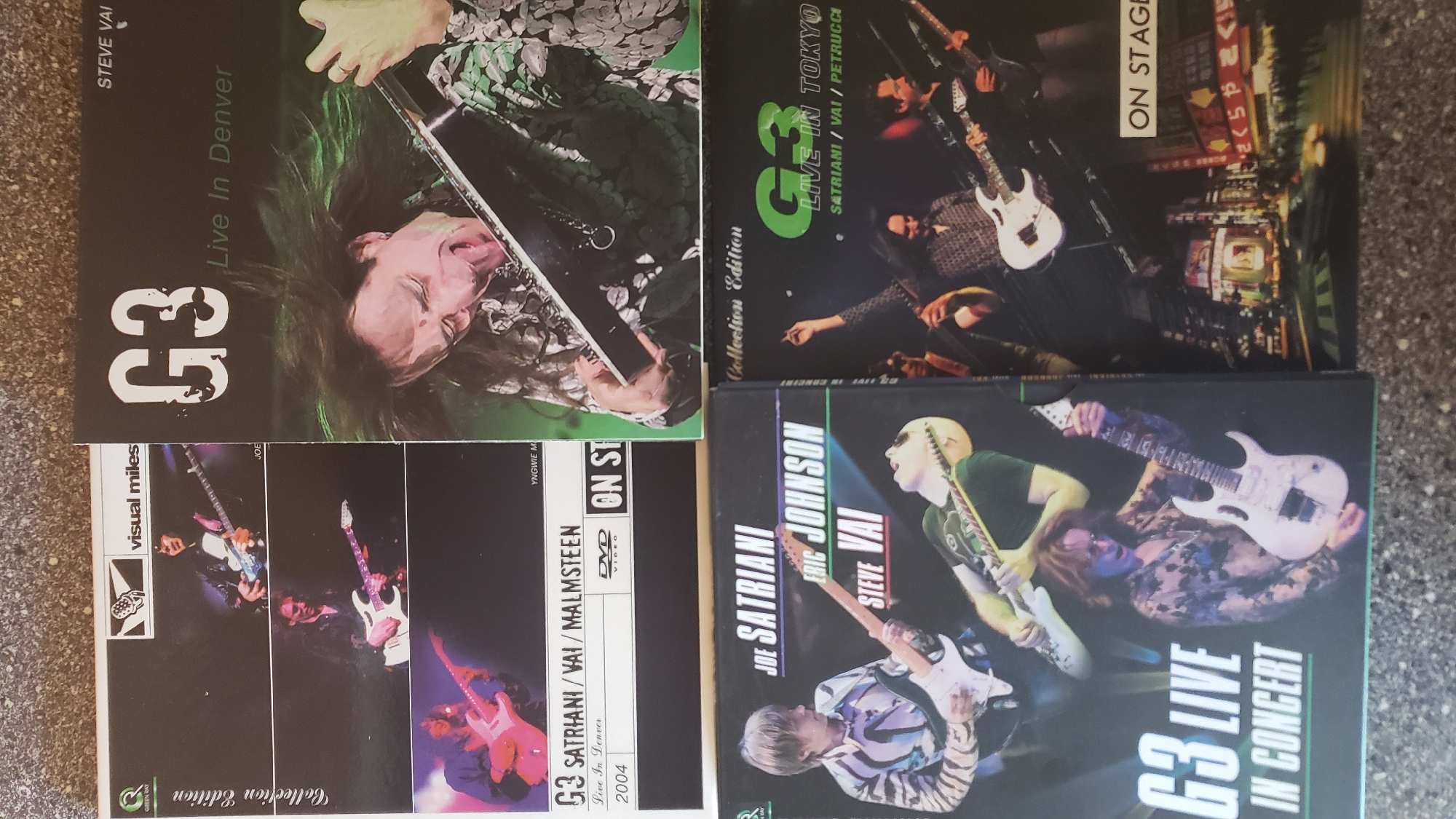 The image displays three DVD covers arranged on a textured surface. The leftmost DVD, titled "G3 SATRIANI / VAI / MALMSTEEN Live In Denver 2004," features three separate images of guitarists on its cover. The middle DVD, "G3 LIVE IN TOKYO," shows a photo of three guitarists performing on stage with a cityscape in the background. The rightmost DVD, also titled "G3 LIVE IN CONCERT," displays images of guitarists Joe Satriani, Eric Johnson, and Steve Vai. There are no discernible landmarks or contextual clues that would indicate a specific location within the city of Prešov, Slovakia. The scene is static and consists solely of the DVD covers.