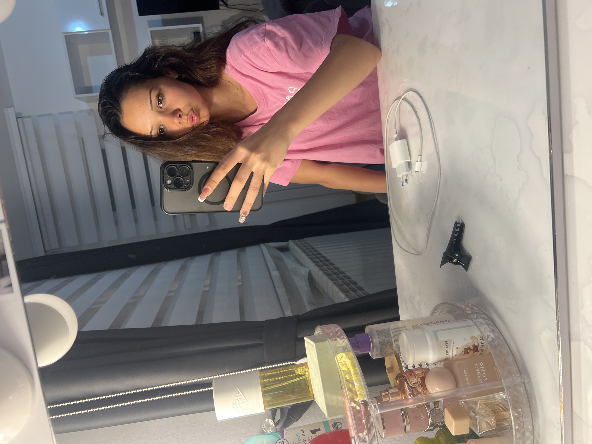 A young woman is captured in a mirror selfie, oriented vertically, inside what appears to be a room. She has long, dark brown hair and is wearing a pink t-shirt with white text that partially reads "COLLEGE". Her lips are slightly pursed in a "pouty" expression, and her gaze is directed towards the camera of her smartphone. She holds a dark-colored smartphone, possibly an iPhone, with multiple camera lenses, in her right hand. Her nails are manicured with a white french tip design, and a ring with a small gem is visible on her ring finger.
The mirror she is using appears to be part of a vanity setup, as indicated by the rounded white lights visible on the left side of the frame. On the white surface of the vanity table to her right, a white USB wall charger and cable are visible. Further to the right and at the bottom of the frame, there's a clear, multi-tiered organizer filled with various bottles, likely cosmetics, perfumes, and lotions. Some text on the bottles is partially visible but largely unreadable due to reflections and angle. A small black hair clip is also seen on the vanity surface.
The reflection in the mirror behind the woman shows horizontal blinds or shutters, suggesting a window is present in the room. Some shelving units are also reflected in the background. The overall lighting suggests an indoor setting, likely using artificial light from the vanity.