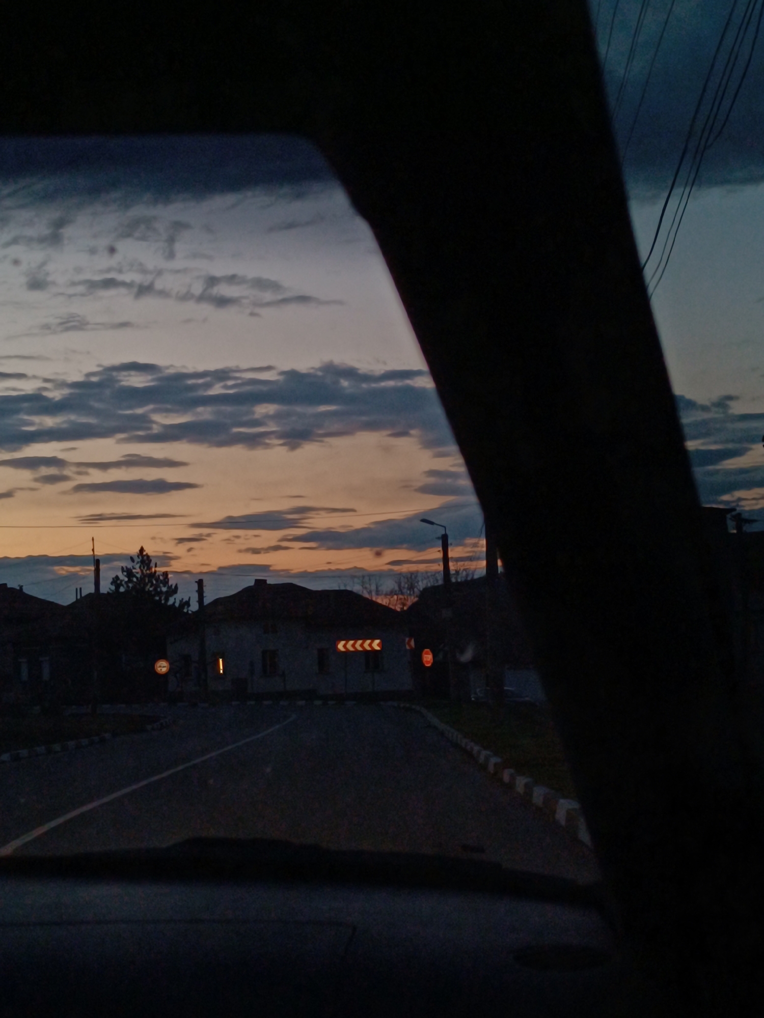Captured from within a vehicle, the image presents a view of a street scene at dusk in an unidentified city. The sky is a blend of blues and oranges, layered with clouds. The street is lined with buildings, featuring lit signage and a solitary illuminated window in one of the structures. There is a utility pole visible near the buildings. The road surface is dark, marked by a white line. A curb of alternating white and gray segments runs alongside the road. The composition is partly obscured by a dark frame, possibly a car window.