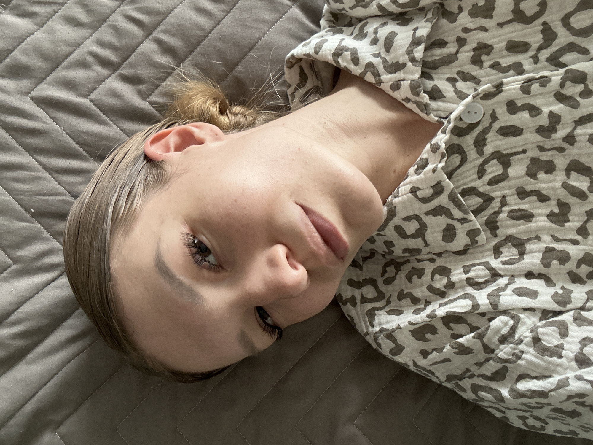 A young woman with blonde hair pulled back into a bun lies on a quilted gray bed. She is wearing a button-up shirt with a leopard print. Her head is turned to the side, and she is looking directly at the camera. Her face is smooth with a few freckles and she has long eyelashes. The lighting suggests the photograph was taken indoors during the daytime. The image does not contain any information that would allow for localization to the city of Chaikovsky, Russia.