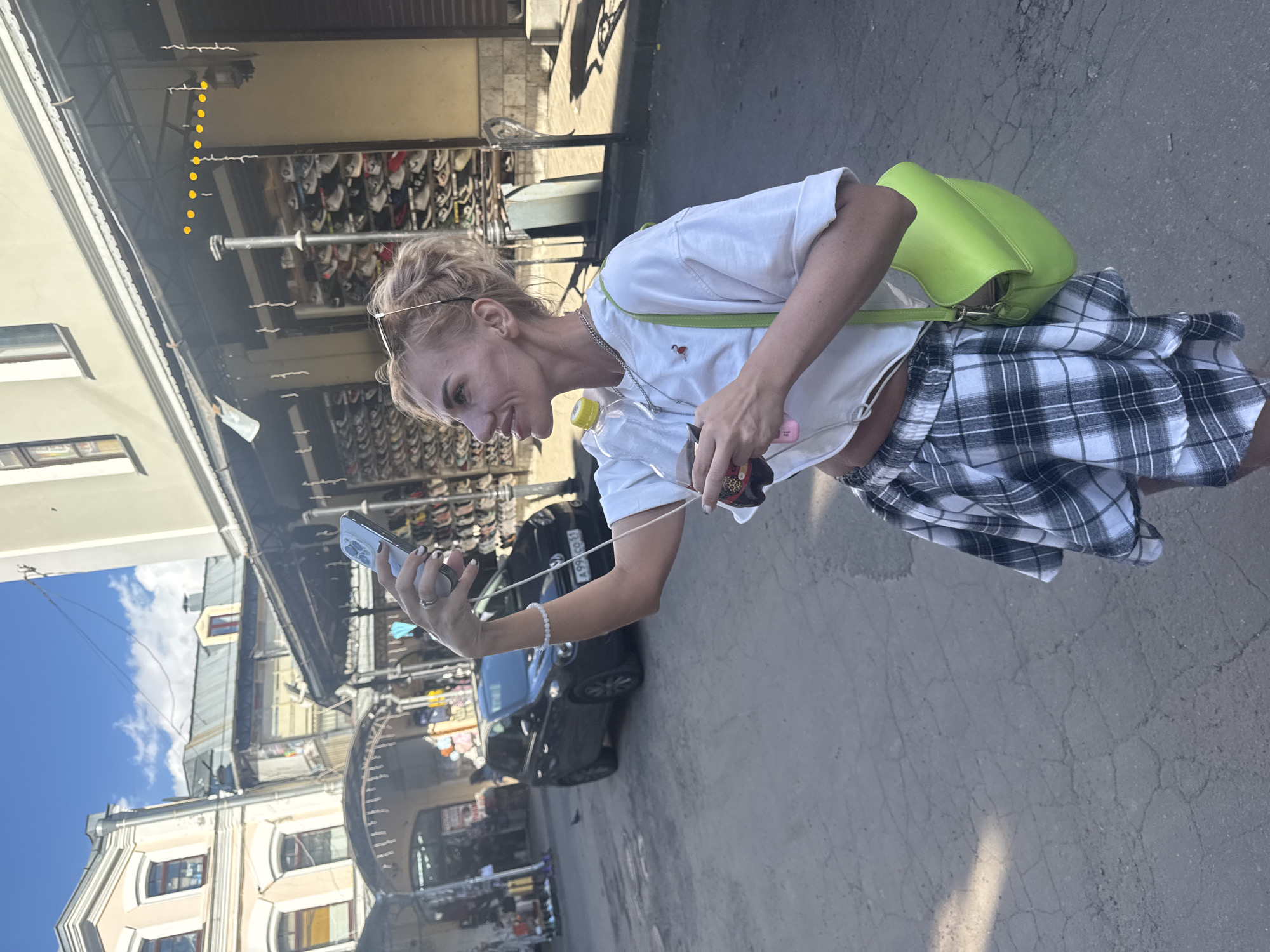 A blonde woman is taking a selfie in an outdoor market on a sunny day. She is wearing a white crop top, a plaid skirt, and a neon green shoulder bag. She is holding a phone in her right hand and a drink in her left. The market appears to be in a city, with buildings and stalls visible in the background. There is a dark car parked on the street behind her. The ground is paved with asphalt and shows signs of wear. Based on the architecture and the presence of a dark car with what appears to be a Russian license plate, the location is likely Saint Petersburg, Russia.