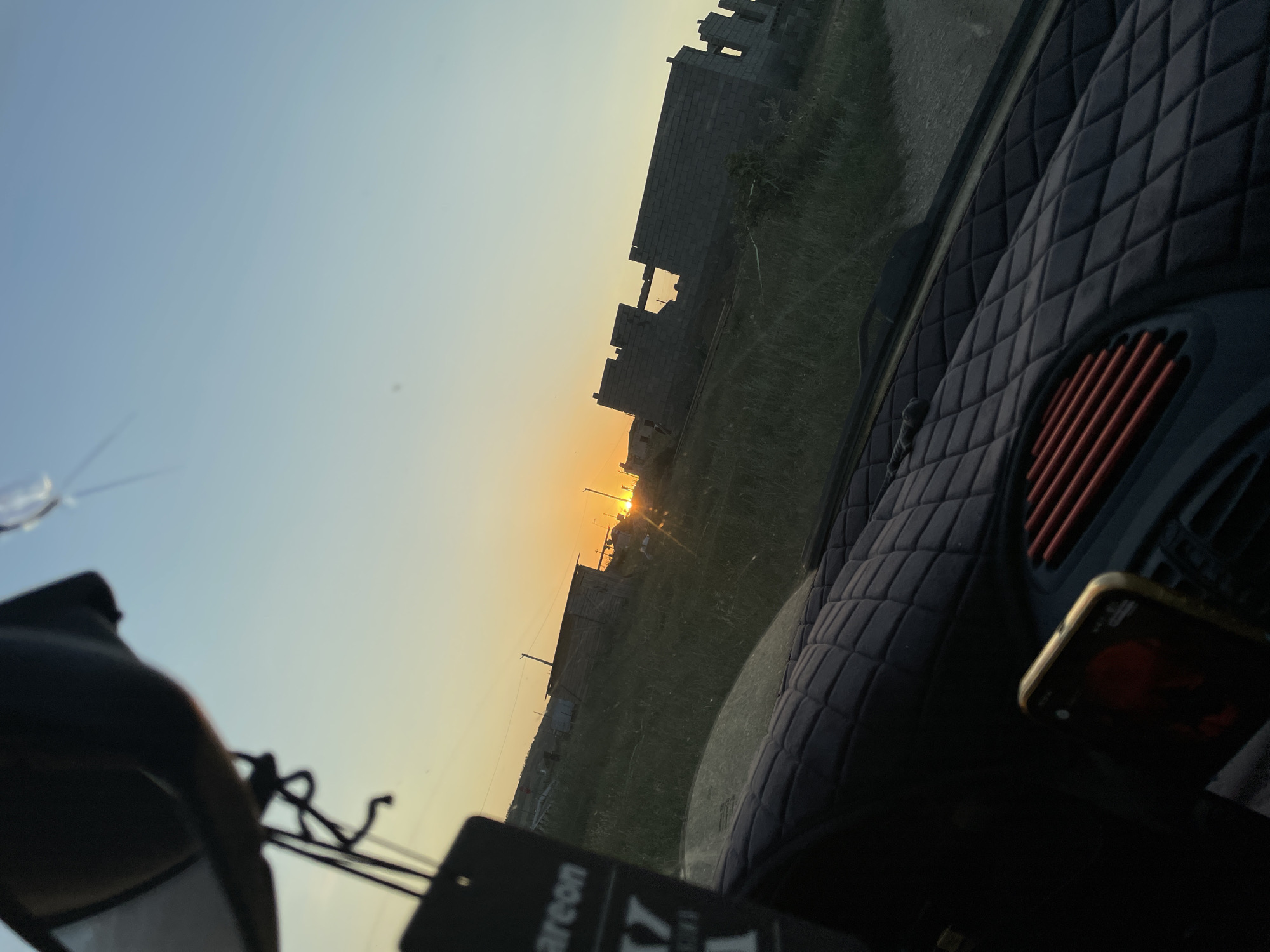 The media file depicts a view from inside a vehicle looking outwards during what appears to be sunset. The scene is set in a semi-urban or rural area, with unfinished brick buildings suggesting construction or a developing neighborhood. The sky is a gradient of pale blue to a warm orange near the horizon, indicative of the golden hour. Green grass and some vegetation are visible in the foreground and around the buildings.
Inside the vehicle, the dashboard is partially visible, featuring a diamond-patterned texture and an air vent with red slats. A smartphone is mounted on the dashboard, displaying a dark screen with some faint elements, possibly notifications or a background image. In the lower left corner of the frame, a dark object, possibly a car accessory or a hanging air freshener, is partially visible. A small tag with visible text hangs from this object.
The overall mood is serene and contemplative, possibly reflecting a moment of travel or observation during the end of the day. The sun is low on the horizon, casting a warm glow and long shadows. The image composition suggests a personal perspective, likely taken by the driver or a passenger. The location context provided, Buynaksk, Russia, implies the setting is within this geographical area.
Visible text on the tag in the lower foreground includes "areon" and a stylized "X7".