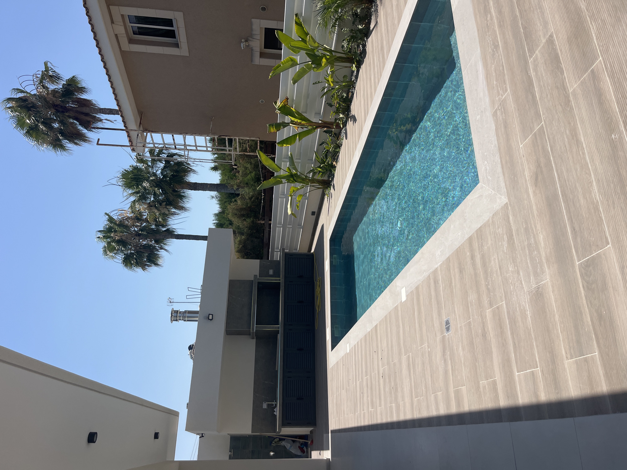This outdoor scene captures a modern residential property in Moni Community, Cyprus, featuring a swimming pool and an outdoor kitchen area. The swimming pool, rectangular in shape, is filled with turquoise water and surrounded by light-colored tiled decking. To the left of the pool, a covered outdoor kitchen is visible, equipped with dark-colored cabinetry, a countertop, and a built-in barbecue grill with a stainless-steel chimney. The background includes a two-story building with a beige facade and a tiled roof, flanked by several tall palm trees. A white horizontal fence separates the property from neighboring areas. The sky is clear and blue, indicating fair weather. The overall impression is one of a newly constructed or renovated outdoor living space designed for leisure.