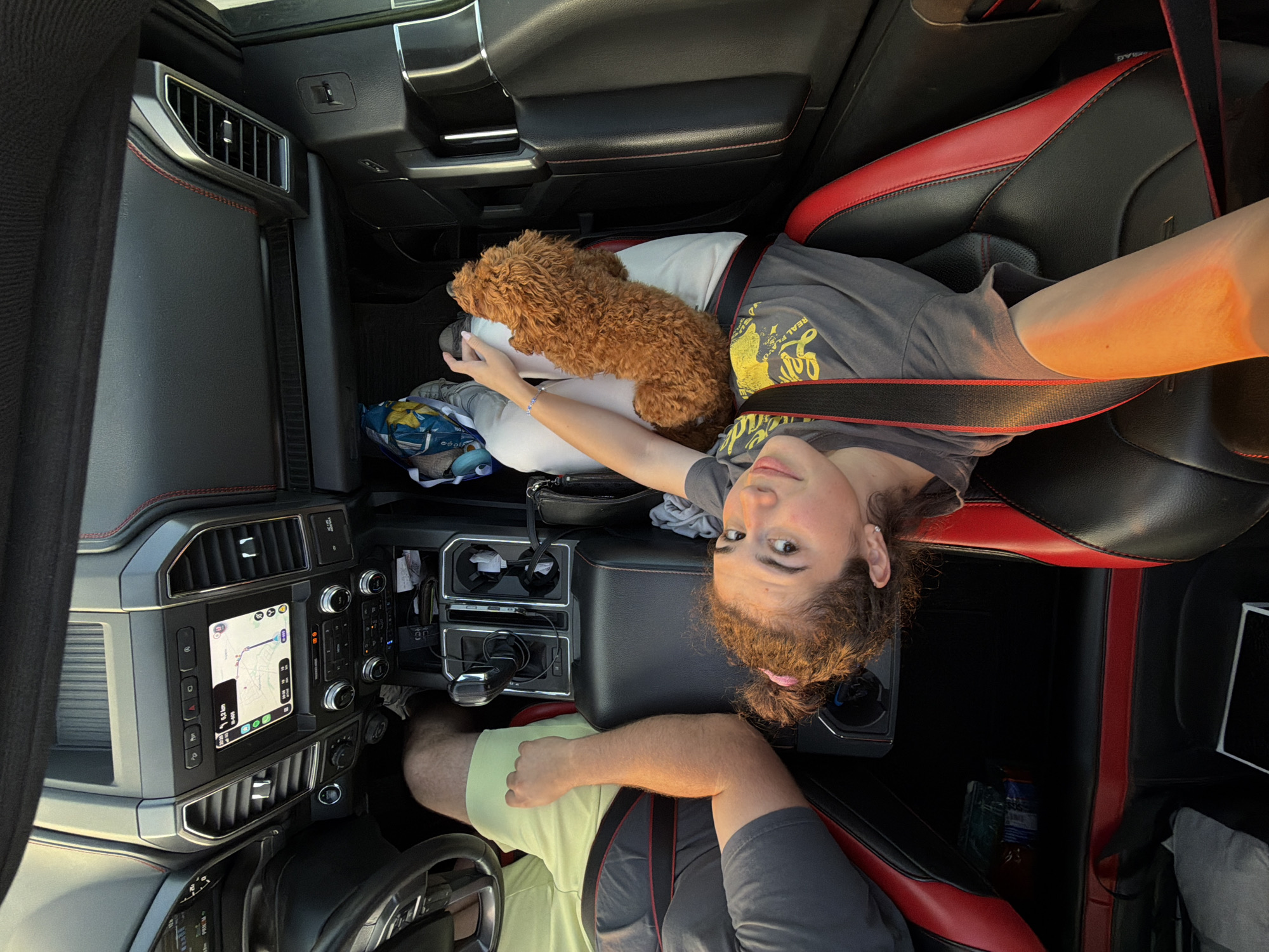 In the interior of a parked vehicle, likely a Ford F-150 based on the dashboard, a young female passenger is lying down across the front seats, with her head near the center console and her body extending towards the passenger seat. She is wearing a grey t-shirt and a seatbelt. To her right, a small, fluffy brown dog is resting on her lap. A male passenger, only his left arm and thigh visible, is seated in the driver's seat, wearing a light green t-shirt. The vehicle's interior features black and red upholstery, a navigation screen displaying a map, and various car controls. Outside the vehicle, sunlight indicates it is daytime. The location is Batanovtsi, Bulgaria.
