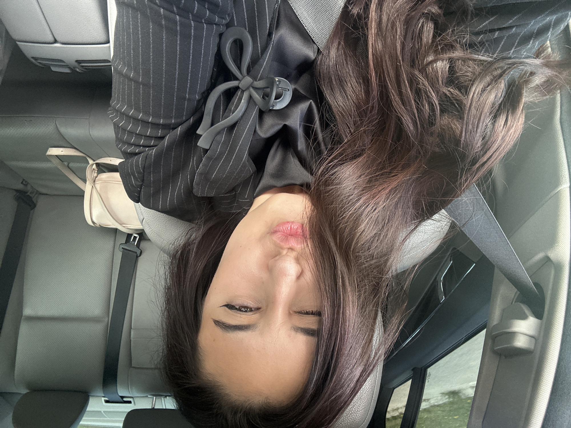 A young woman is in the backseat of a car. She wears a black pinstripe blazer over a black top with a decorative bow. She is wearing a seatbelt. A cream-colored purse is on the seat next to her. She is looking directly at the camera with her lips pursed and one eye partially closed. The location appears to be in Varna, Bulgaria. No further information is available from the image.