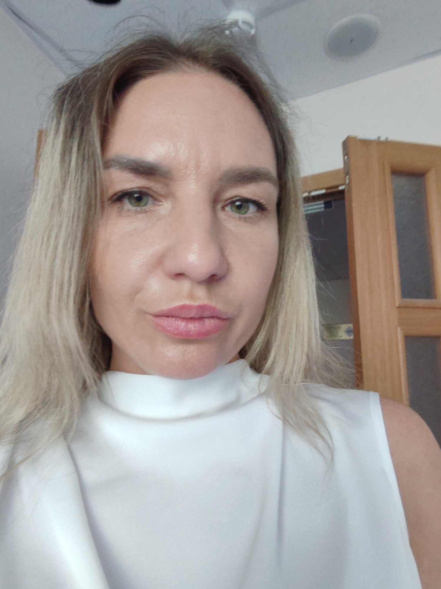 A close-up, waist-up shot shows a fair-skinned woman with blonde hair and green eyes. She is wearing a white sleeveless top with a high neckline. The woman is looking directly at the camera and has her lips pursed. In the background, a wooden door is visible. The setting appears to be indoors, possibly a home or office.