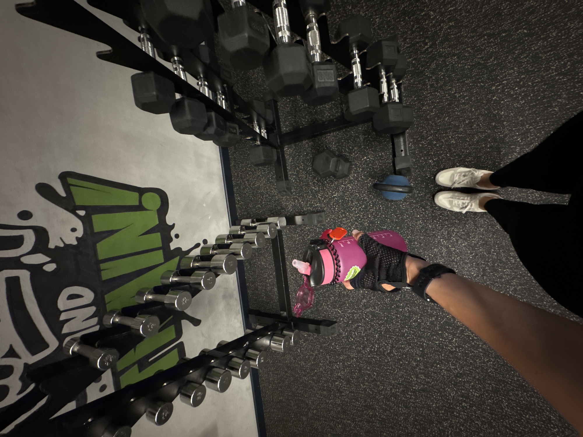 The image shows a person's arm holding a pink water bottle in a gym. There are racks of dumbbells in the background, both chrome and black hexagonal ones. A logo with the words "TRAIN AND TRAIN" is visible on the wall. The floor is a speckled grey rubber mat. The person is wearing black workout gloves and black leggings. White sneakers are visible on their feet. The scene does not provide any information about the city of Sochi, Russia.