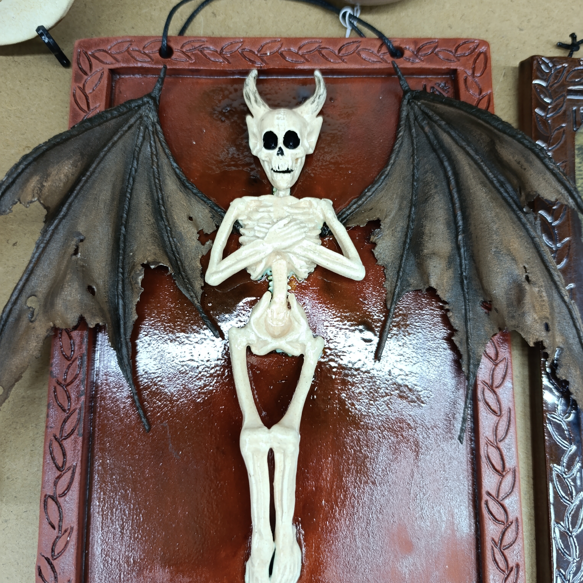 A decorative art piece featuring a winged skeleton with horns is displayed on a brown ceramic tile with a raised leaf-patterned border. The skeleton has its arms crossed over its chest. The wings are dark brown and tattered. The item is hung by a black cord. Another similarly bordered item is partially visible to the right. The photo was taken in the city of Chaikovsky, Russia.