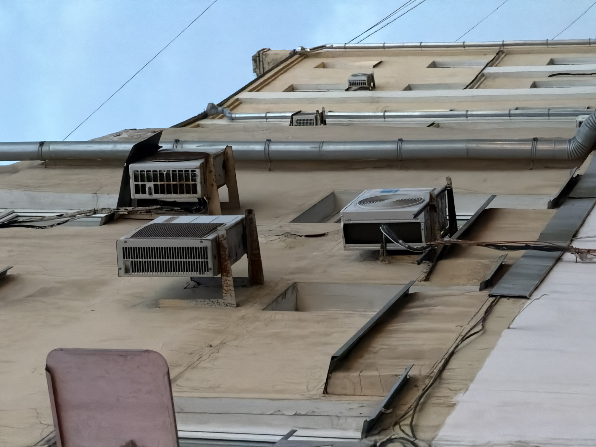 The image shows the exterior wall of a building in an unknown city. The wall is tan and has several air conditioning units attached to it. There are also pipes running along the wall and metal sheets and other objects attached to the wall. The sky is visible in the background.