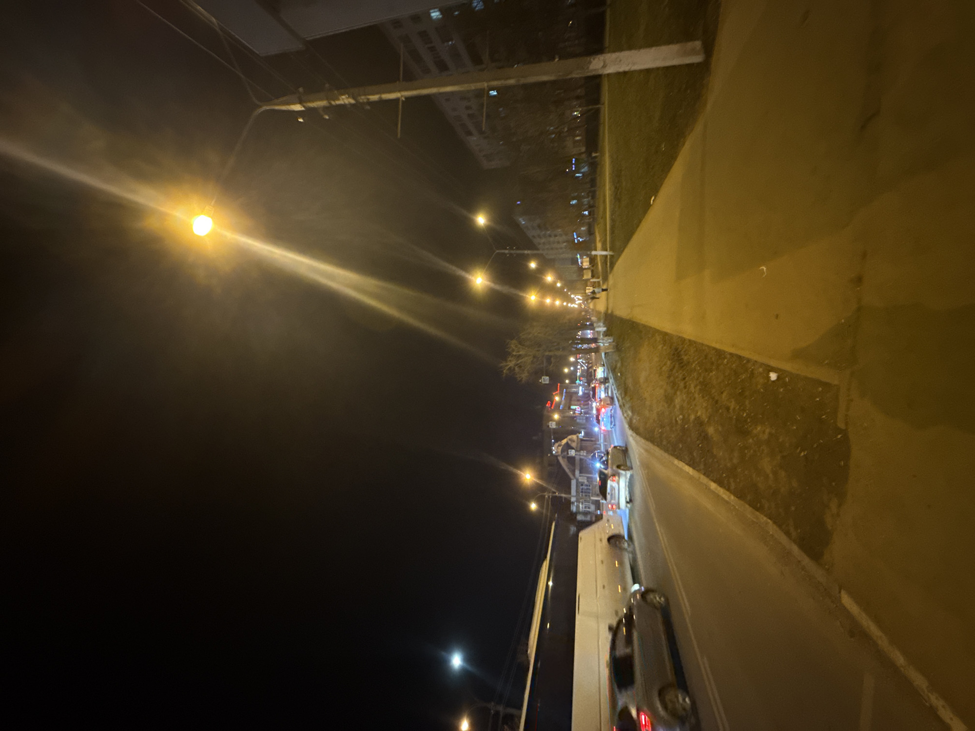 This is a night scene on a street in what appears to be Dmitrov, Russia. The image is taken at ground level, possibly from the street. A long row of streetlights illuminates a road with cars and a sidewalk. Adjacent to the sidewalk is a grassy area. Further back, one can discern buildings along the street and additional street lighting, suggesting a busy city environment. Some sort of construction vehicles can be seen further down the street. The scene is characterized by artificial light from streetlights and vehicle headlights.