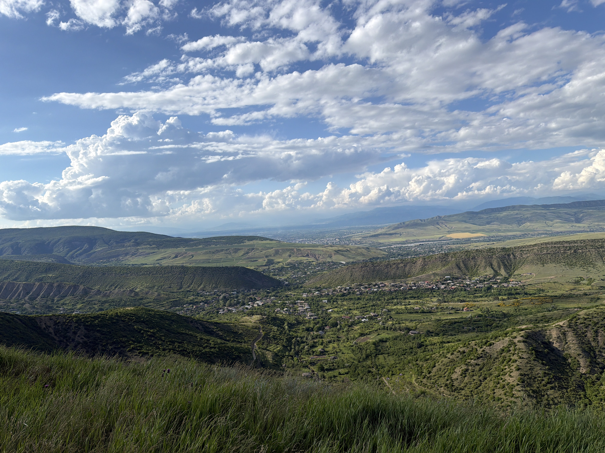 The image captures a panoramic view of a valley landscape in Ateni, Georgia. The scene is dominated by rolling green hills and mountains, with a small village nestled in the valley below. The foreground features tall, green grass that is slightly out of focus, creating a sense of depth. In the mid-ground, the village appears as a collection of buildings with red roofs, surrounded by lush vegetation and scattered trees. A winding dirt path can be seen leading towards the village. The background shows a series of layered hills and mountains fading into the distance, with a hint of a river or body of water.
The sky is a vibrant blue, filled with a variety of clouds, ranging from wispy cirrus to larger, more dramatic cumulus formations. The sunlight filtering through the clouds suggests it is daytime, likely late morning or early afternoon. The weather appears to be fair, with clear skies and fluffy clouds contributing to a picturesque and serene atmosphere. There is no discernible activity or event taking place; the image captures a static, natural landscape. No people, objects, or text are clearly visible within the frame. The overall impression is one of natural beauty and tranquility, showcasing the rural landscape of Ateni, Georgia.