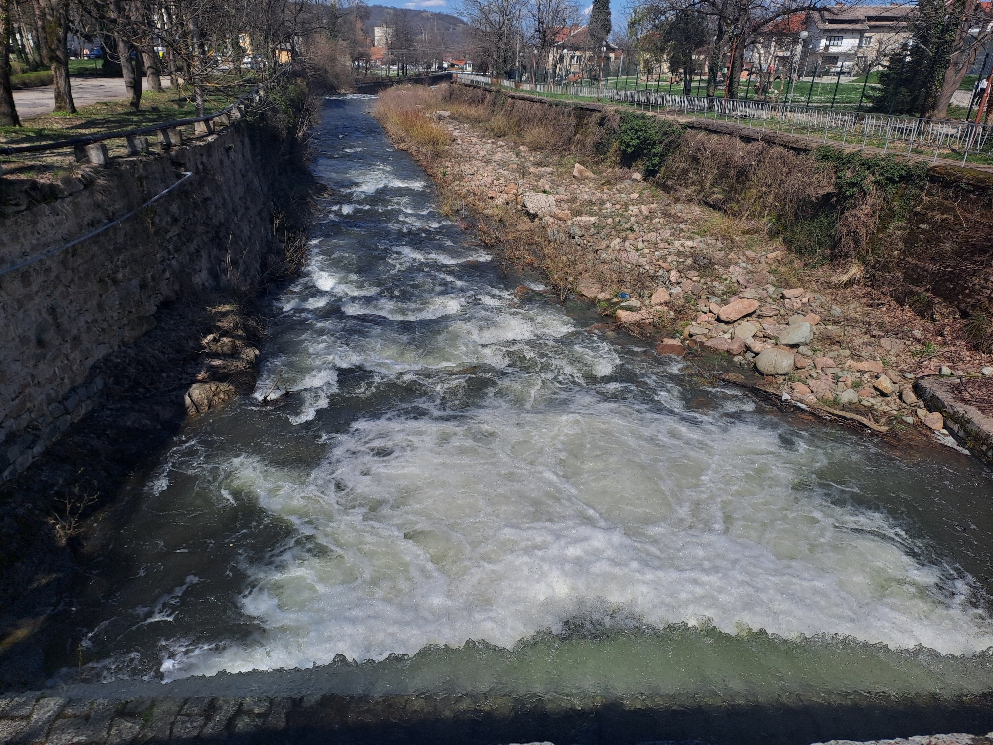 The image shows a river flowing through a channelized section in the city of Varshets, Bulgaria. The river is turbulent with white water visible. The channel is lined on both sides with stone or concrete walls and embankments. The right bank has a rocky area with exposed soil and vegetation. A fence is visible along the top of the right bank. Trees line the sides of the river. In the distance, buildings and a tower can be seen. The scene appears to be in an urban or suburban setting on a sunny day. No people are directly visible in the image.