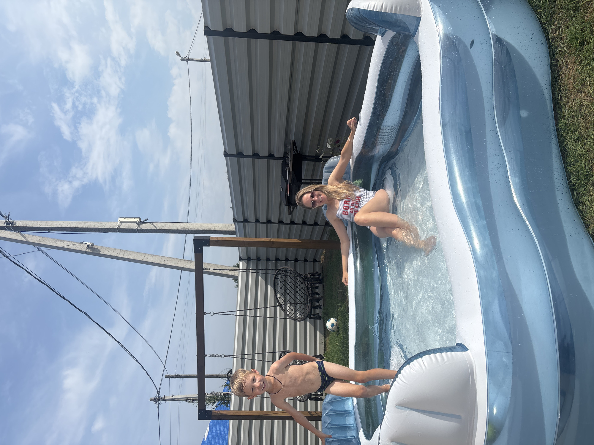 In an outdoor scene on a sunny day, a young boy and a woman are enjoying an inflatable swimming pool. The boy, wearing dark swim trunks with a yellow pattern, stands in the shallow end of the pool, smiling at the camera. A woman in a red and white striped swimsuit is positioned in the deeper section of the pool, her arm extended upwards. The pool is partially shaded by a corrugated metal roof structure supported by wooden beams. A black woven hanging chair is visible in the background, along with a white soccer ball on the grass. Power lines and concrete poles crisscross the sky, suggesting a residential or semi-rural setting. The weather appears clear and bright, indicated by the blue sky and scattered white clouds. The overall mood seems relaxed and joyful, as the two subjects are engaged in a leisure activity on a warm day.