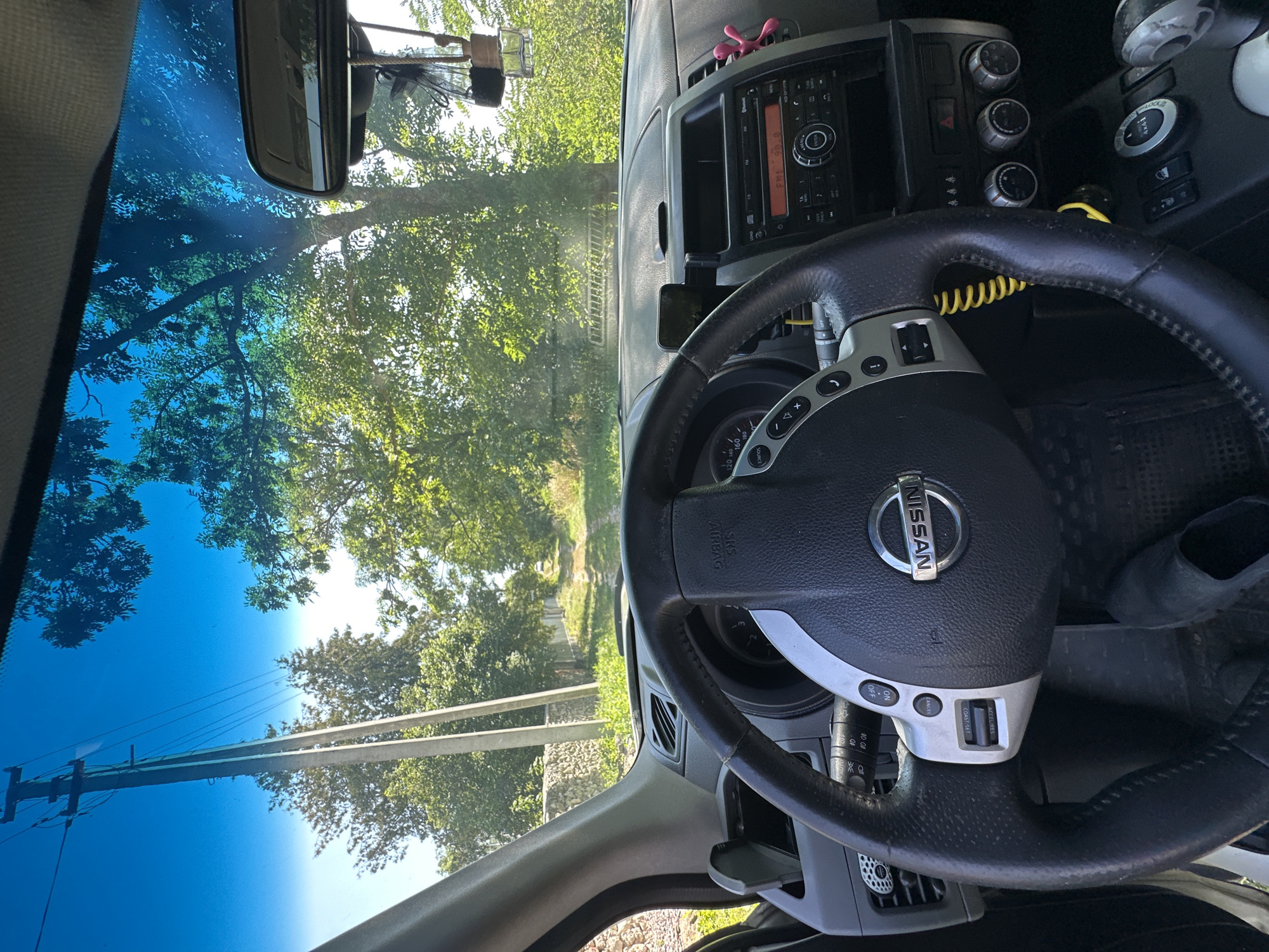 The image shows the interior of a car looking out through the windshield at a rural road. The car's steering wheel, dashboard, and center console are visible. Outside, a dirt road lined with trees and a power pole is seen under a clear blue sky. The car appears to be parked or driving slowly on a sunny day. The location is specified as Makiv, Ukraine.