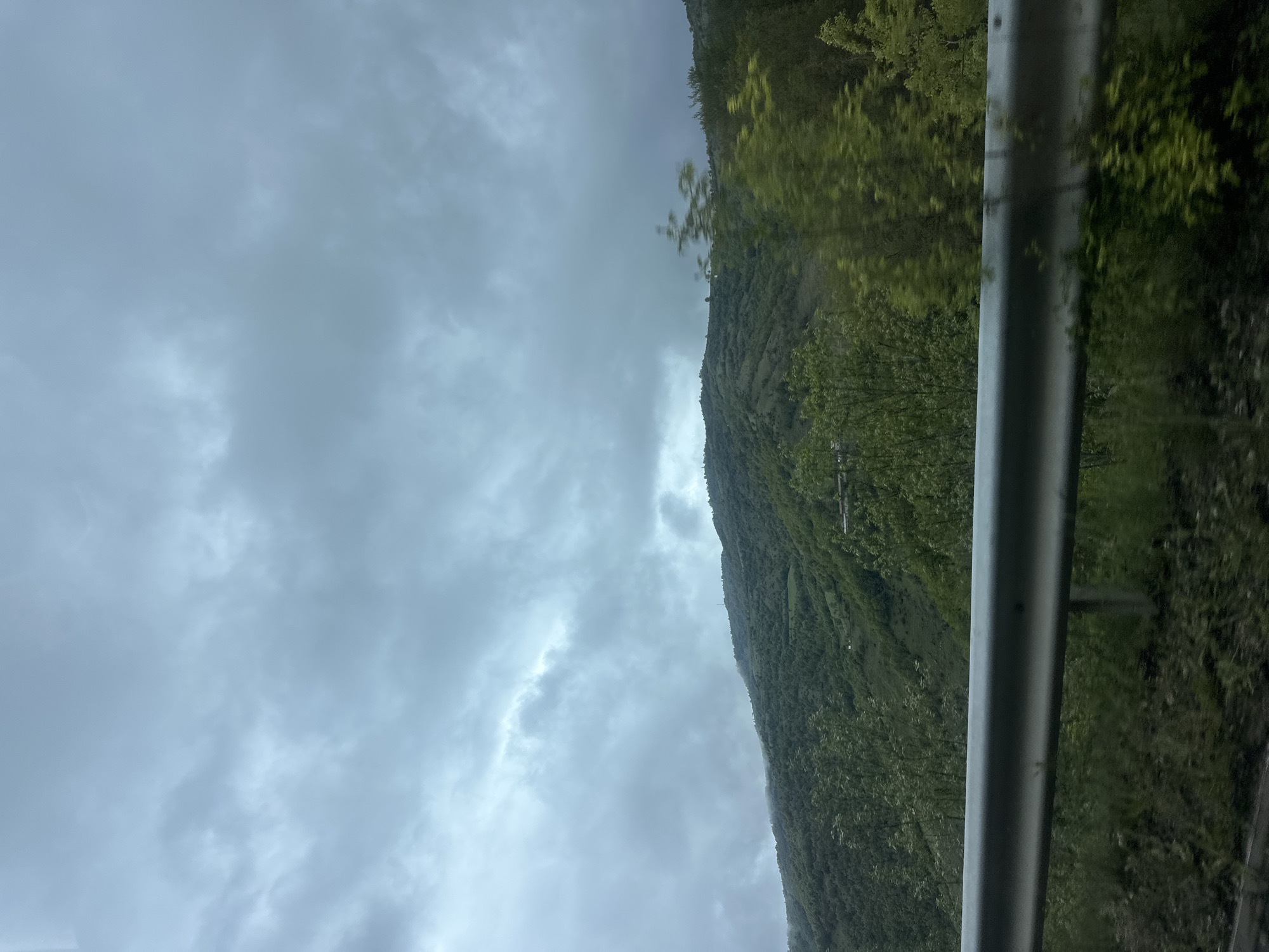 The image shows a landscape taken from a moving vehicle, likely a car, as evidenced by the guardrail on the right side of the frame. The left side of the image is dominated by a cloudy, overcast sky. To the right, a mountain covered in dense green vegetation rises sharply. The location is potentially near Liaskovo, Bulgaria, based on the request, although specific landmarks to confirm this are absent. No people or distinct subjects besides the natural environment are identifiable. The action is implied movement of the vehicle taking the photo.