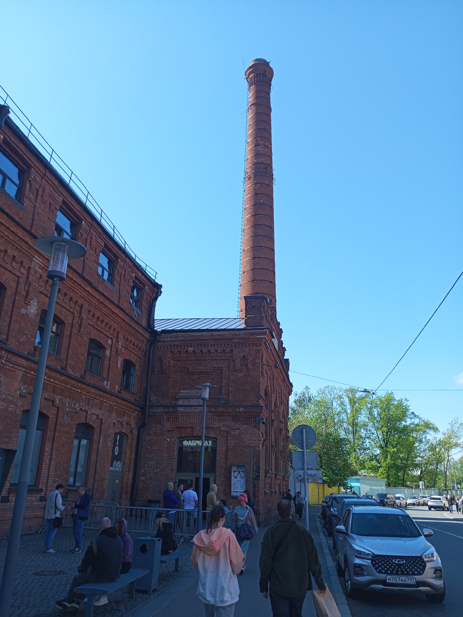The image depicts the Red Brick Building with a distinctive smokestack and is identified as "Fabrika A3 Factory" in Saint Petersburg, Russia. People are seen both inside and outside of the building. A car is parked along the side of the street adjacent to the brick building. The sky is bright blue and trees are in the background, suggesting it is daytime.