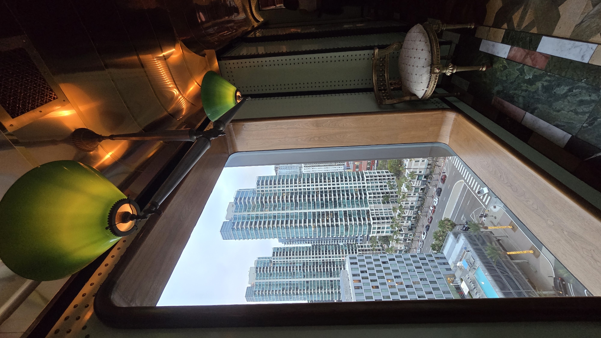 The image shows an interior view from a window of a building, looking out at a city skyline and street scene. On the interior side, there are two green pendant lamps hanging from the ceiling, which appears to be made of polished metal and has industrial-style rivets. The window frame is made of wood. To the right of the window, there is an ornate, gold-colored chair with a patterned seat. The floor is a mosaic of different colored stone tiles, primarily green, white, and pink.
The view outside the window reveals a dense urban environment. Several tall, modern glass-and-steel skyscrapers dominate the skyline. In the foreground, there is a street with cars, crosswalks, and palm trees lining the sidewalks. A swimming pool and an adjacent building with a grid-like facade are also visible. The sky is overcast.
Given the architectural style of the buildings, particularly the prominent skyscrapers that are characteristic of the downtown area, this scene is likely located in the Marina District or Columbia District of San Diego, California, United States of America.