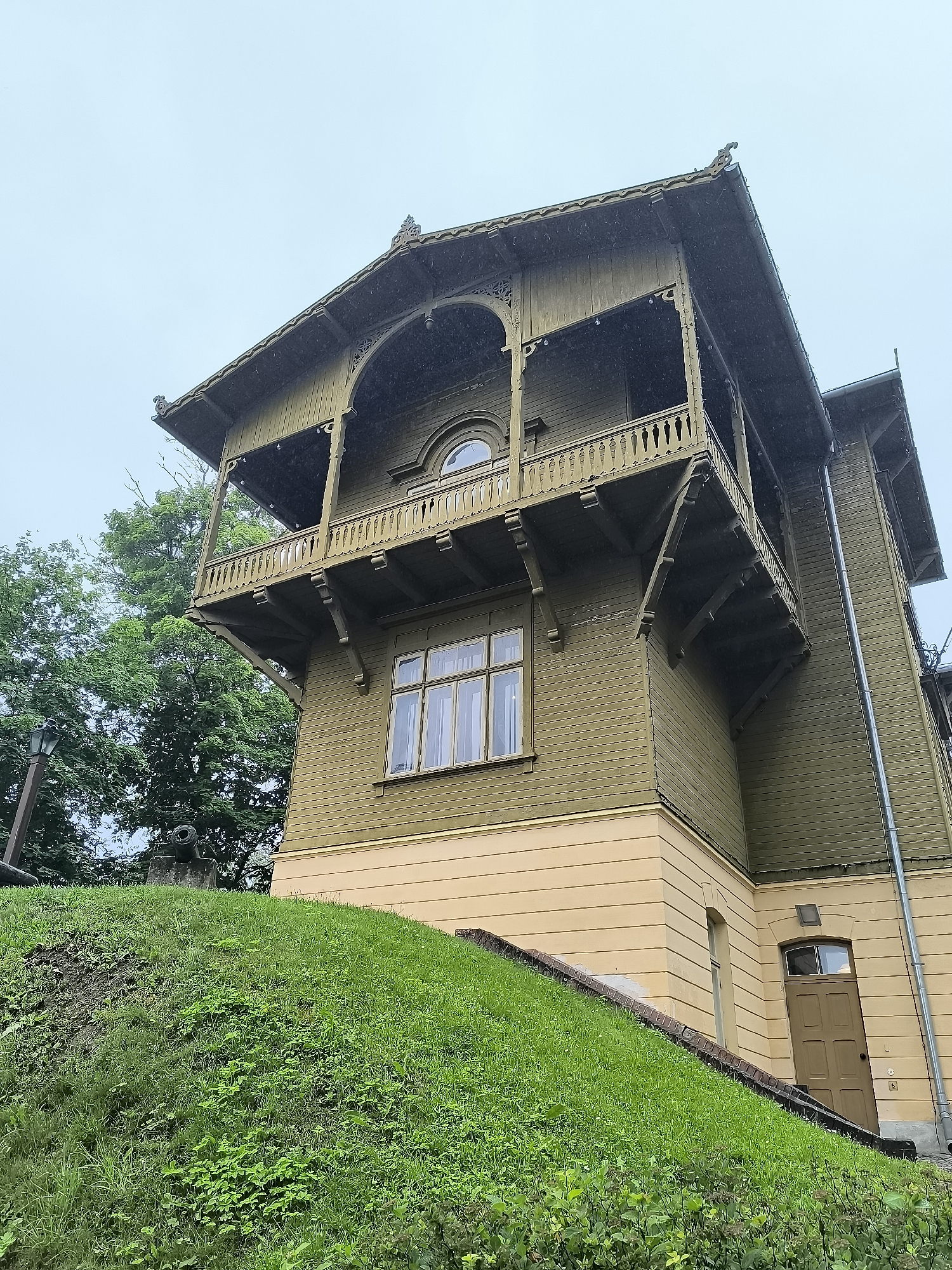 The image shows a multi-story wooden house with green siding and a yellow base, located on a grassy incline. The house features a prominent balcony with ornate railings and decorative eaves. A window with multiple panes is visible on the lower level, and an arched window is present on the upper level. To the left of the house, there are trees and a lamppost. At the base of the grassy incline, there appears to be a historical cannon. The sky is overcast, suggesting it might be raining or foggy, with visible water droplets in the air. This property is known as the Rendas Manor in Kuldīga, Latvia.