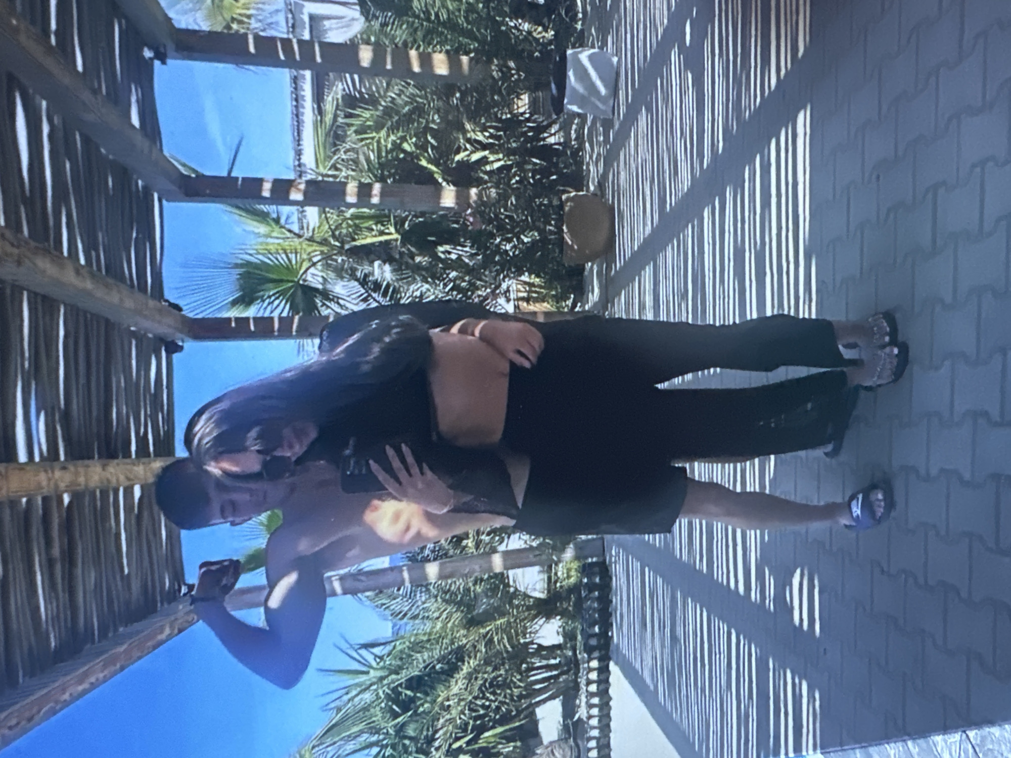 The image shows a young couple posing for a selfie outdoors. The man, shirtless and flexing his bicep, stands behind the woman. The woman, wearing a black crop top and pants, holds a smartphone to take the picture. They are standing under a wooden pergola, with palm trees and a bright blue sky visible in the background. The ground is paved with interlocking stones, and strong shadows from the pergola create a striped pattern. The location is not identifiable as Sofia, Bulgaria.