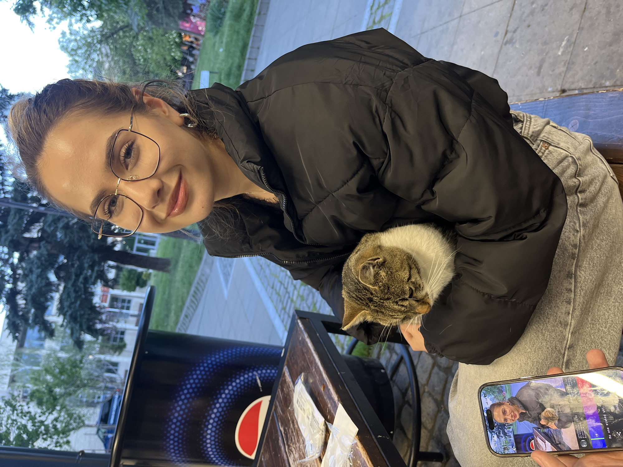 A young woman in a black puffer jacket and glasses is holding a tabby and white cat. She is sitting at an outdoor table with a dark wooden top and a black Pepsi-branded base. The woman is smiling towards the camera. Another hand holds a smartphone displaying a video of the same woman and cat in what appears to be the same location. The background shows a street scene with trees, buildings, and a light-colored pedestrian walkway. Based on the elements visible and the prompt, the location is in the city of Blagoevgrad, Bulgaria.