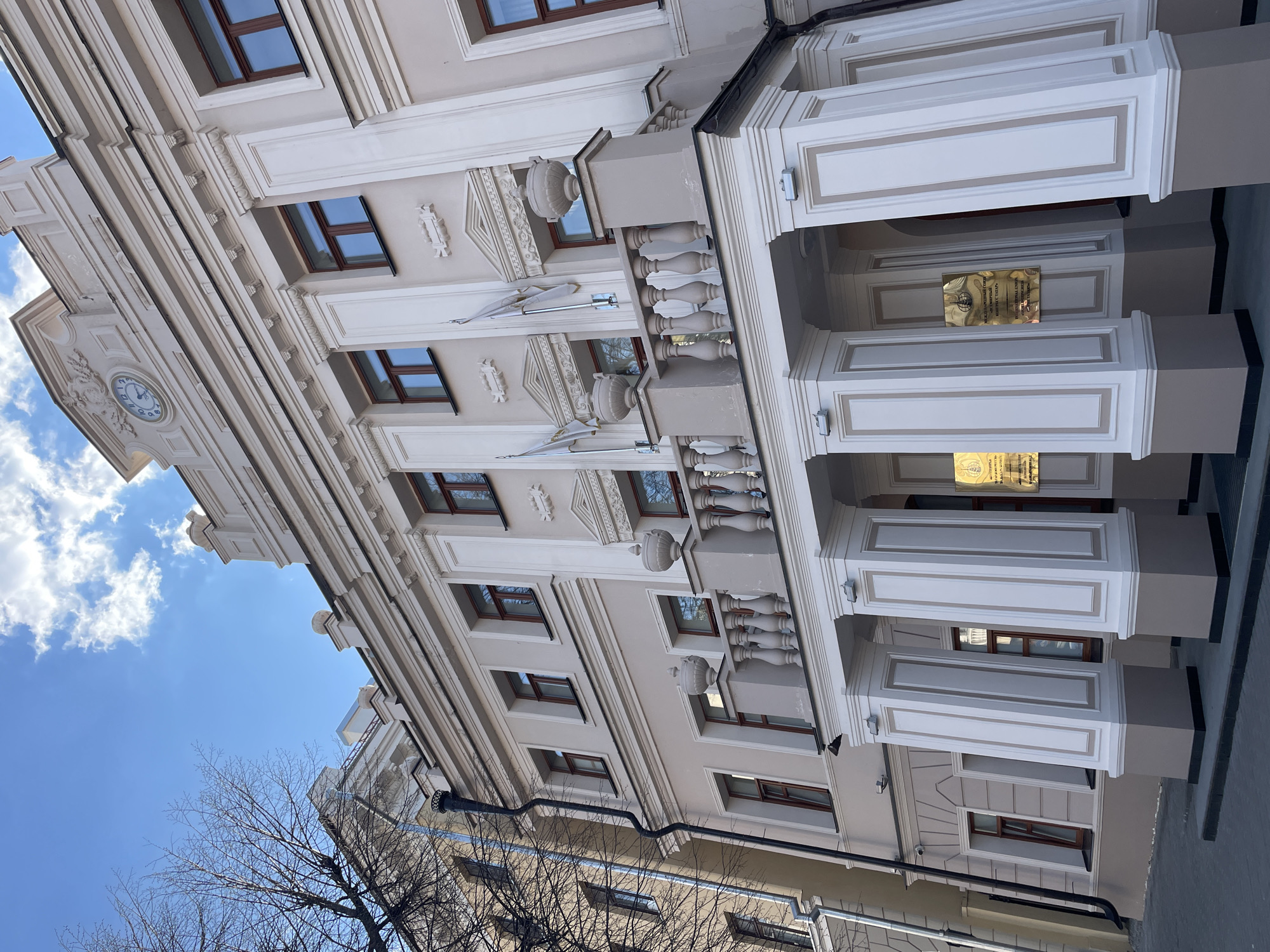 The image shows a building in Kazan, Russia. The building has a beige facade with white trim and brown window frames. There's a clock on the top left of the facade and a balcony with decorative stone elements, including urns and balustrades, near the entrance. There are plaques mounted on the columns flanking the entrance. Bare tree branches are visible in the lower left corner, and part of another building is visible in the lower portion of the image. The sky is visible in the upper left corner, with some clouds. The image is taken from a low angle, looking upward.