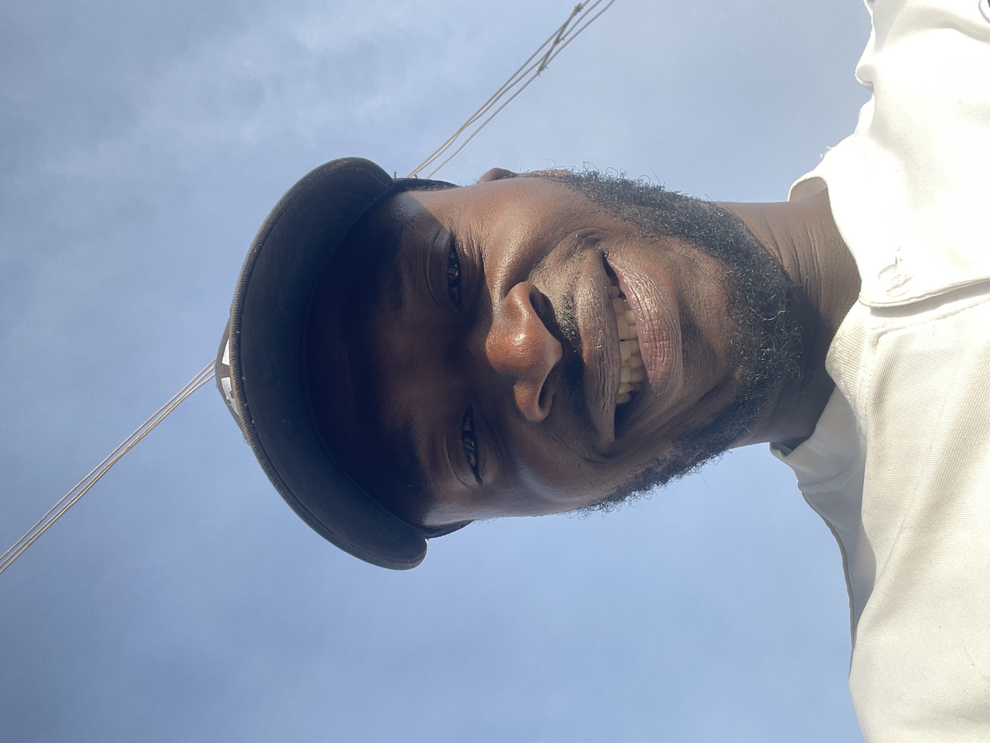 The image is a close-up, low-angle portrait of a dark-skinned man smiling broadly, revealing his teeth. He has a beard and mustache, and his expression conveys cheerfulness. He is wearing a dark cap, visible on the left side of his head, and a light-colored, possibly white, collared shirt is partially visible on the far right of the frame.
The photograph is oriented sideways, with the man's head appearing on the right side of the frame, taking up a significant portion of the image. The background is predominantly a bright, clear blue sky with some very faint clouds, indicating an outdoor setting during daytime. Several thin, light-colored wires or cables traverse the upper left quadrant of the sky. Bright sunlight illuminates the man's face, highlighting his skin tone and features. The perspective is from below, looking up at the subject. The scene is set in Jos, Nigeria.
There is no visible text in the image.