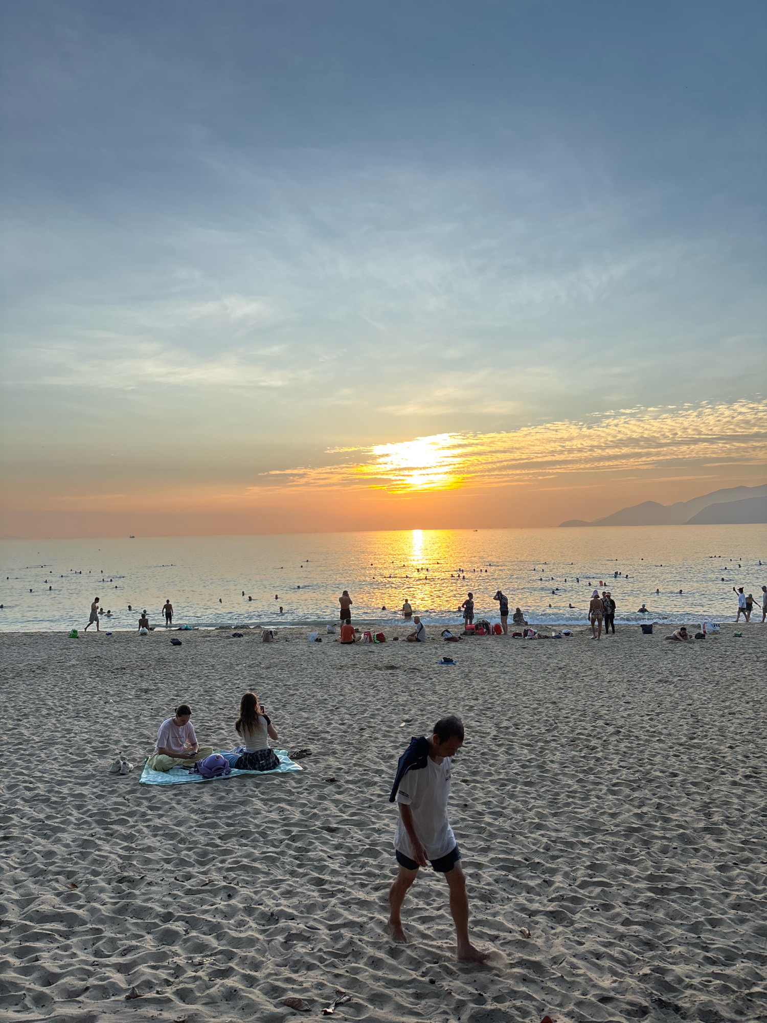 This is a photograph of a beach in Nha Trang, Vietnam, during sunset. The sun is low on the horizon, casting a warm orange glow across the water and sky. There are many people on the beach and in the water, some swimming, others relaxing on towels. The sand is a light beige color, and there are a few scattered leaves and shells. In the foreground, a man in a white t-shirt and dark shorts walks across the sand. In the middle ground, two women sit on a blanket, one on her phone, the other looking at her phone. The sea is calm, with gentle waves lapping at the shore. The sky is a mix of blue and orange, with a few wispy clouds. In the distance, the silhouettes of mountains can be seen against the horizon.