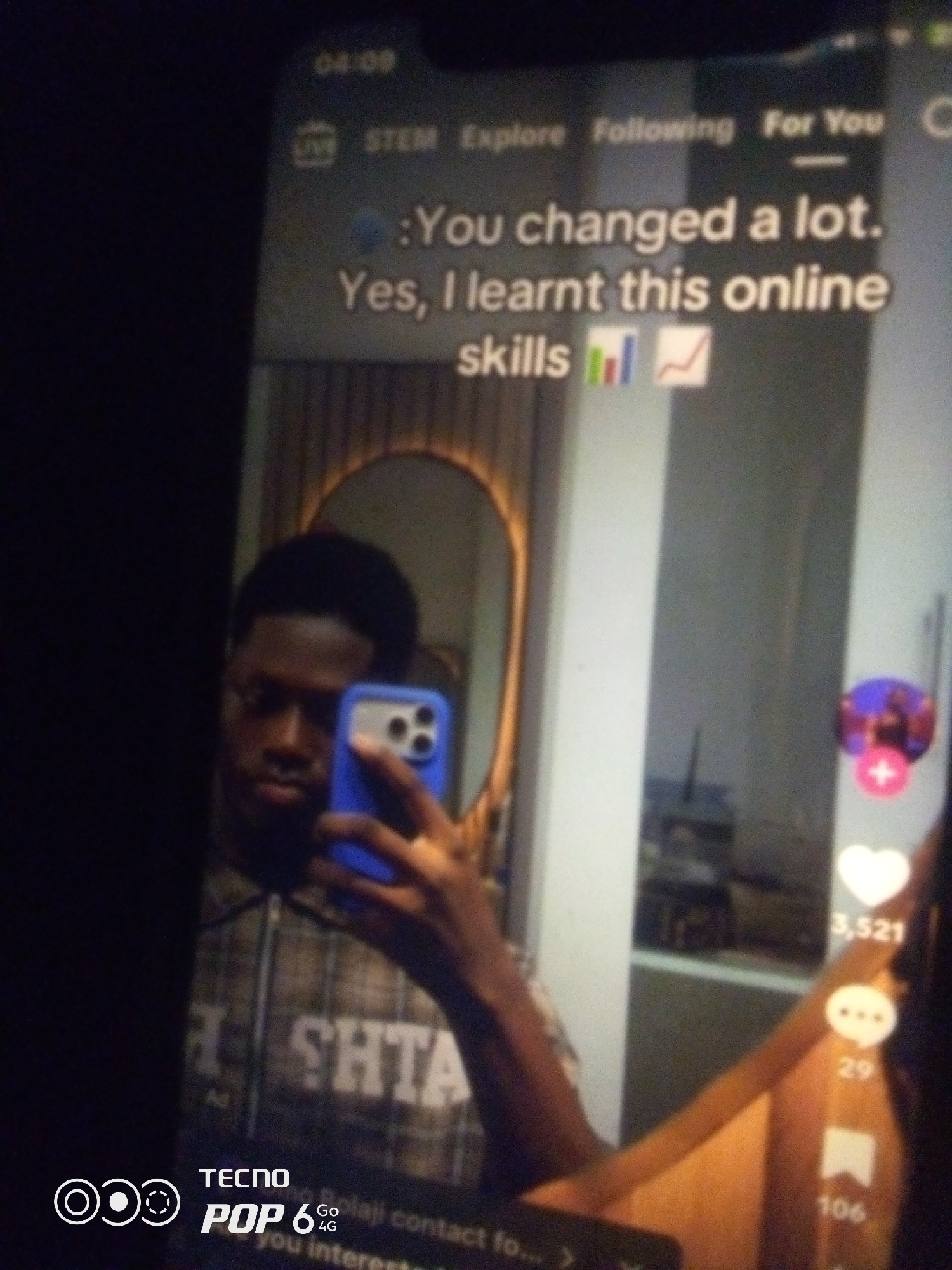 The image is a close-up shot of a person taking a selfie in a room. The person is holding a blue-cased smartphone in their right hand, which is angled to capture their reflection in a mirror. The reflection shows the person's face, a patterned shirt, and a hint of the room's interior. On the smartphone screen, text is visible, including "You changed a lot." and "Yes, I learnt this online skills". There are also social media interface elements like icons for likes, comments, and shares, with associated numbers. The overall lighting suggests an indoor environment, possibly at night or in a dimly lit space. The image does not contain any specific visual cues that would indicate a location in Howayi, Nigeria.
