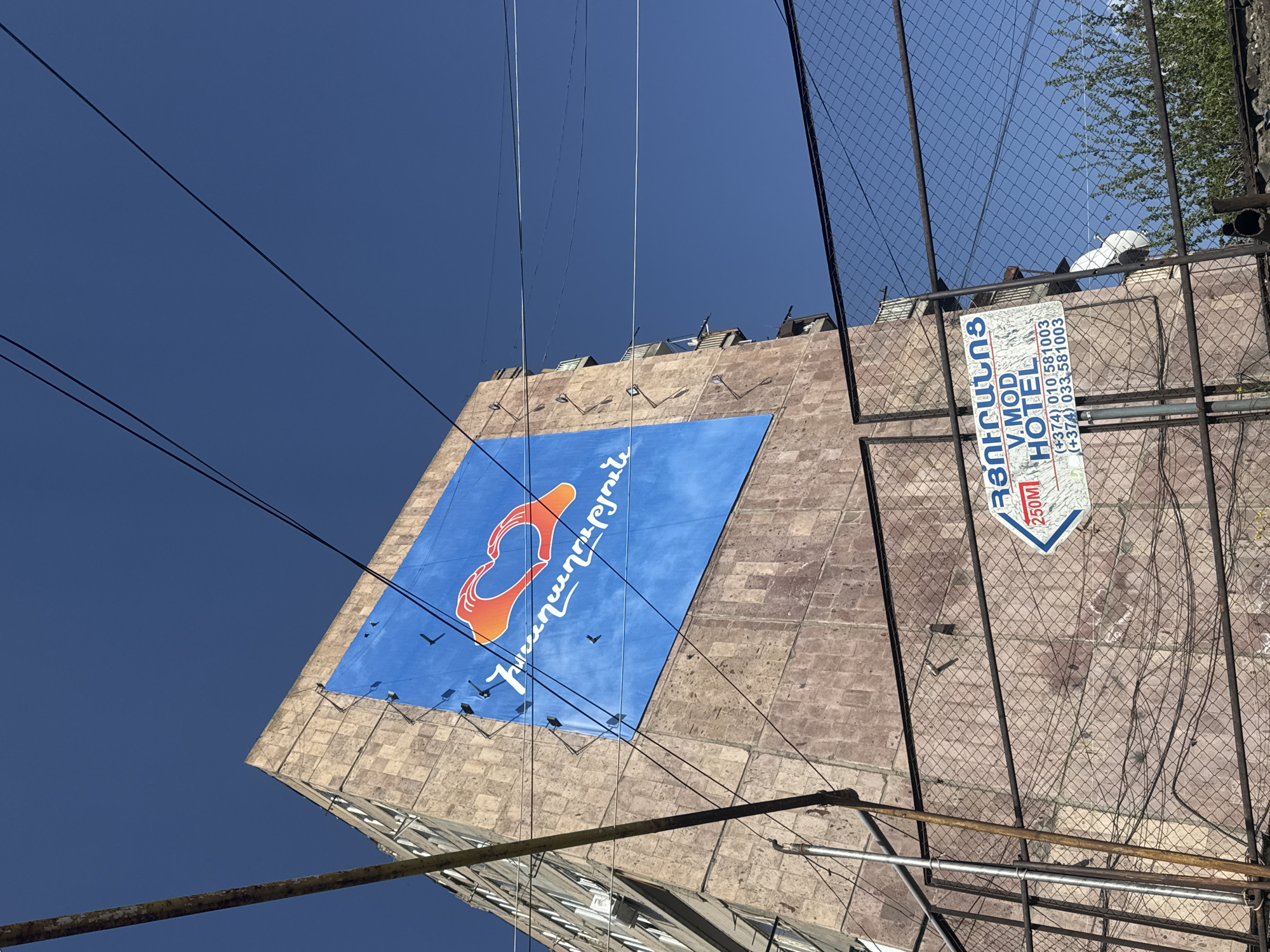 The image shows a building in Yerevan, Armenia, with a large blue sign featuring an orange logo and Armenian script. The building's facade is composed of stone blocks. Above the sign are multiple spotlights. Power lines crisscross the foreground against a clear blue sky. A metal fence and a sign for the "V. MOD Hotel" are visible on the right side of the image, along with what appears to be a satellite dish and some trees. The photograph is taken from a low angle, looking upwards.