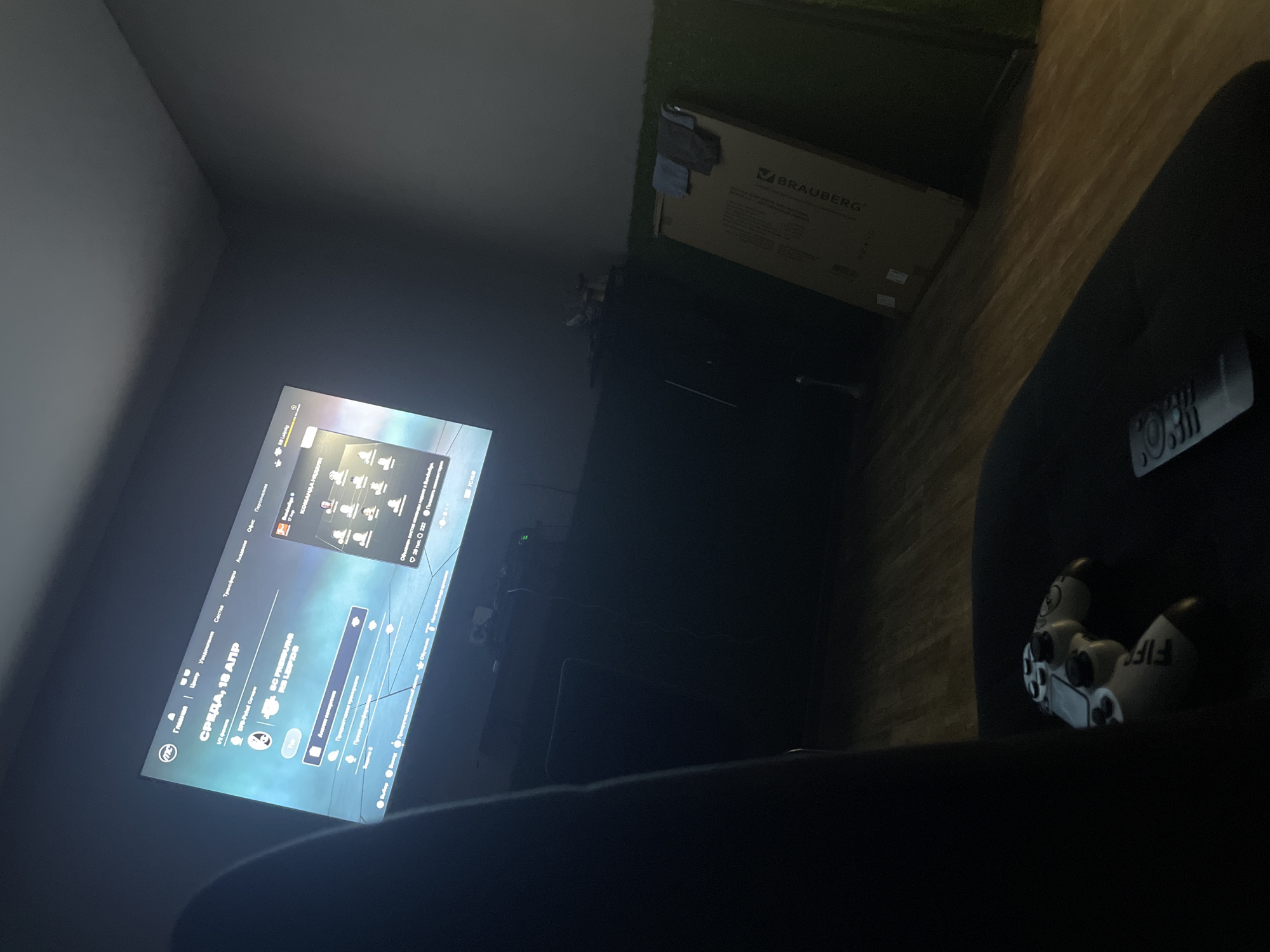 A dimly lit room features a large television screen displaying a soccer video game interface, likely FIFA. The screen shows team information, player avatars, and menu options in Russian. A black gaming chair is partially visible in the foreground, with a white and gray video game controller resting on its seat. A silver remote control is also present on the seat. In the background, a dark cabinet or entertainment unit is visible, with various electronic devices and cables. To the right of the screen, a large cardboard box with the "BRAUBERG" logo is leaning against a wall, and a piece of green artificial turf is visible near the floor. The floor appears to be made of wooden planks. The scene is consistent with a home entertainment setup, possibly in Nevinnomyssk, Russia, given the Russian language interface on the screen.