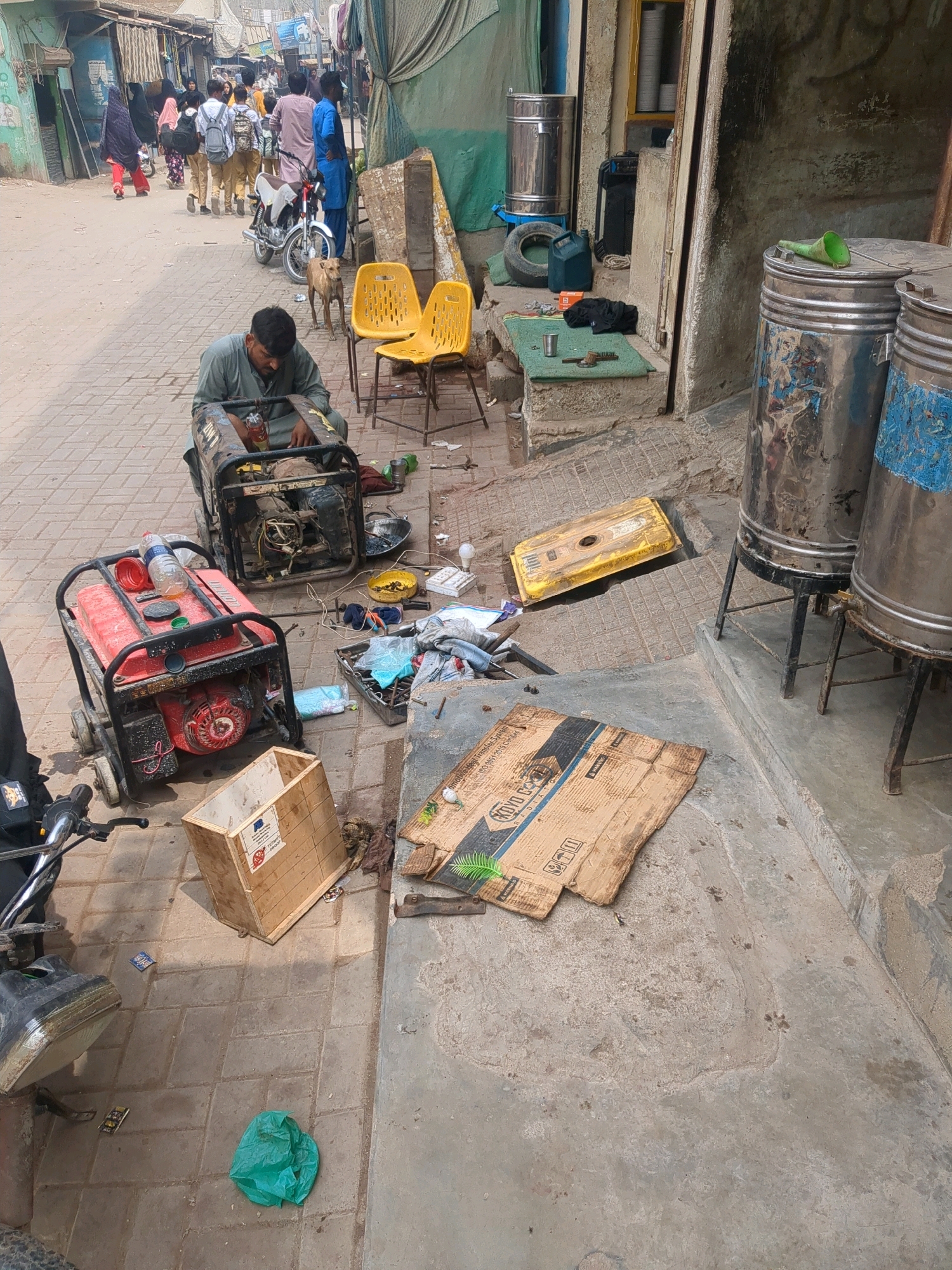A street scene in Karachi Division, Pakistan, shows a man in a green shirt working on a portable generator on a paved street. In the foreground, a red generator is also visible, along with a wooden crate, a green plastic bag, and various tools and discarded items. In the background, people are walking down the street, some carrying backpacks, and a motorcycle is parked. Two yellow chairs are set up next to the street, and a dog is standing nearby. The buildings lining the street appear to be simple structures, with awnings and exposed wiring. The overall impression is one of a bustling, informal urban environment.