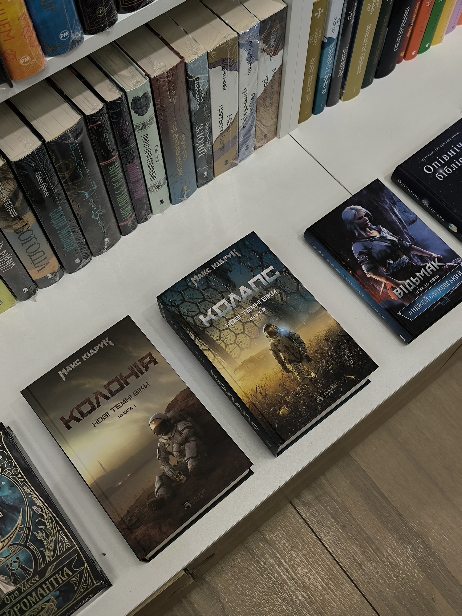 The image shows a bookshelf in Vinnytsia, Ukraine. The books are arranged on a white shelf. The spines of the books are visible, and some are in Ukrainian. The books "КОЛОНІЯ" and "КОЛАПС" by Макс Кідрук are visible. The Witcher series, translated into Ukrainian, can also be seen. The floor is wooden.