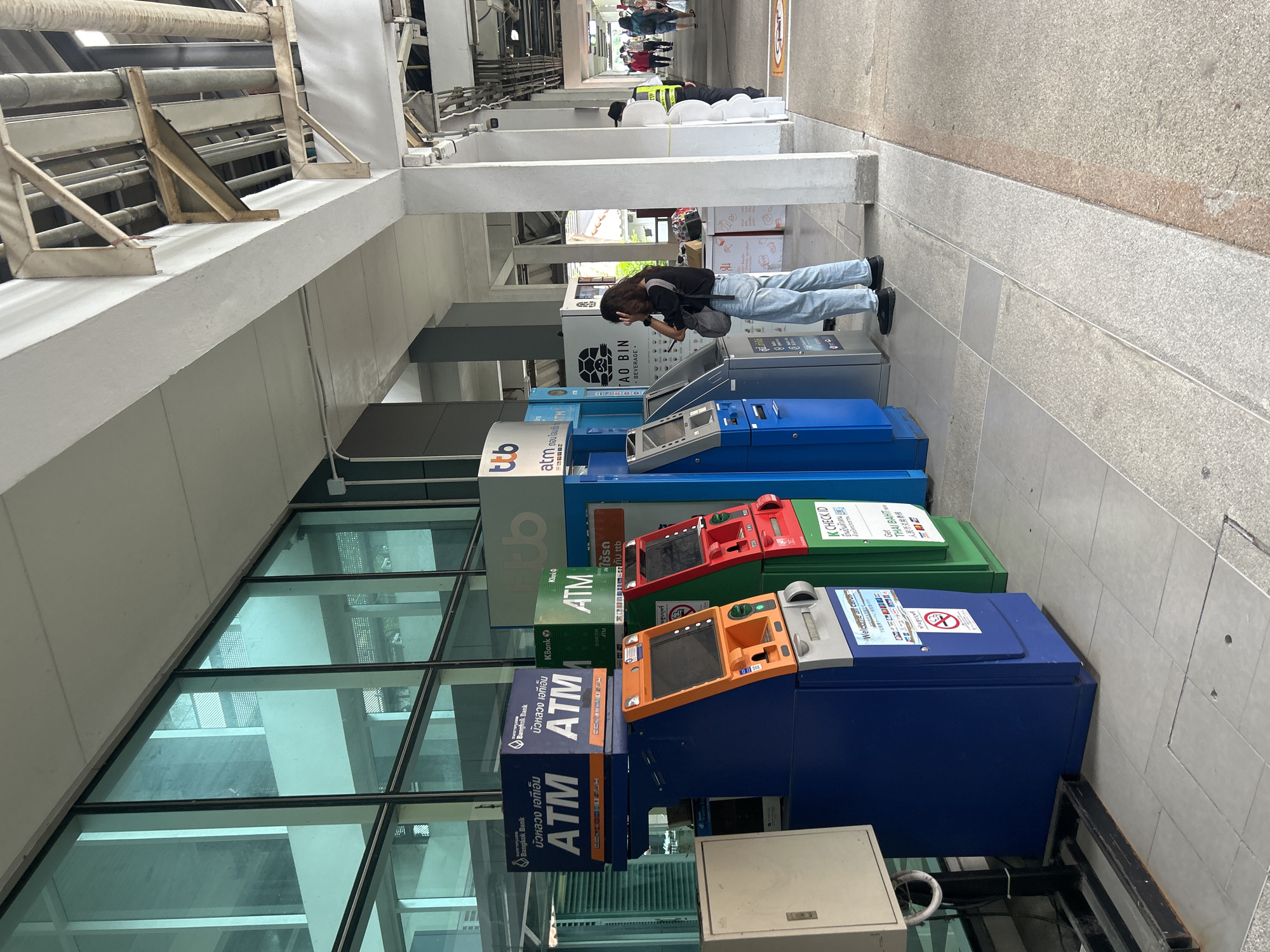 The image depicts a row of ATMs situated outside a modern building with a large glass facade. The ATMs are of various colors including blue, orange, green, and silver. Several are branded with bank names such as Bangkok Bank, ttb, and KBank. A woman with long dark hair, wearing a black t-shirt and light blue jeans, stands in front of one of the ATMs, appearing to be interacting with it. She is carrying a grey shoulder bag. Further down the walkway, there are other individuals, including a person in a yellow vest, and a few people walking in the distance. The environment appears to be an outdoor public space, possibly a transportation hub or a commercial area, with overhead structures and piping visible. The ground is paved with tiles and concrete. The overall scene suggests a typical urban setting in Thailand.