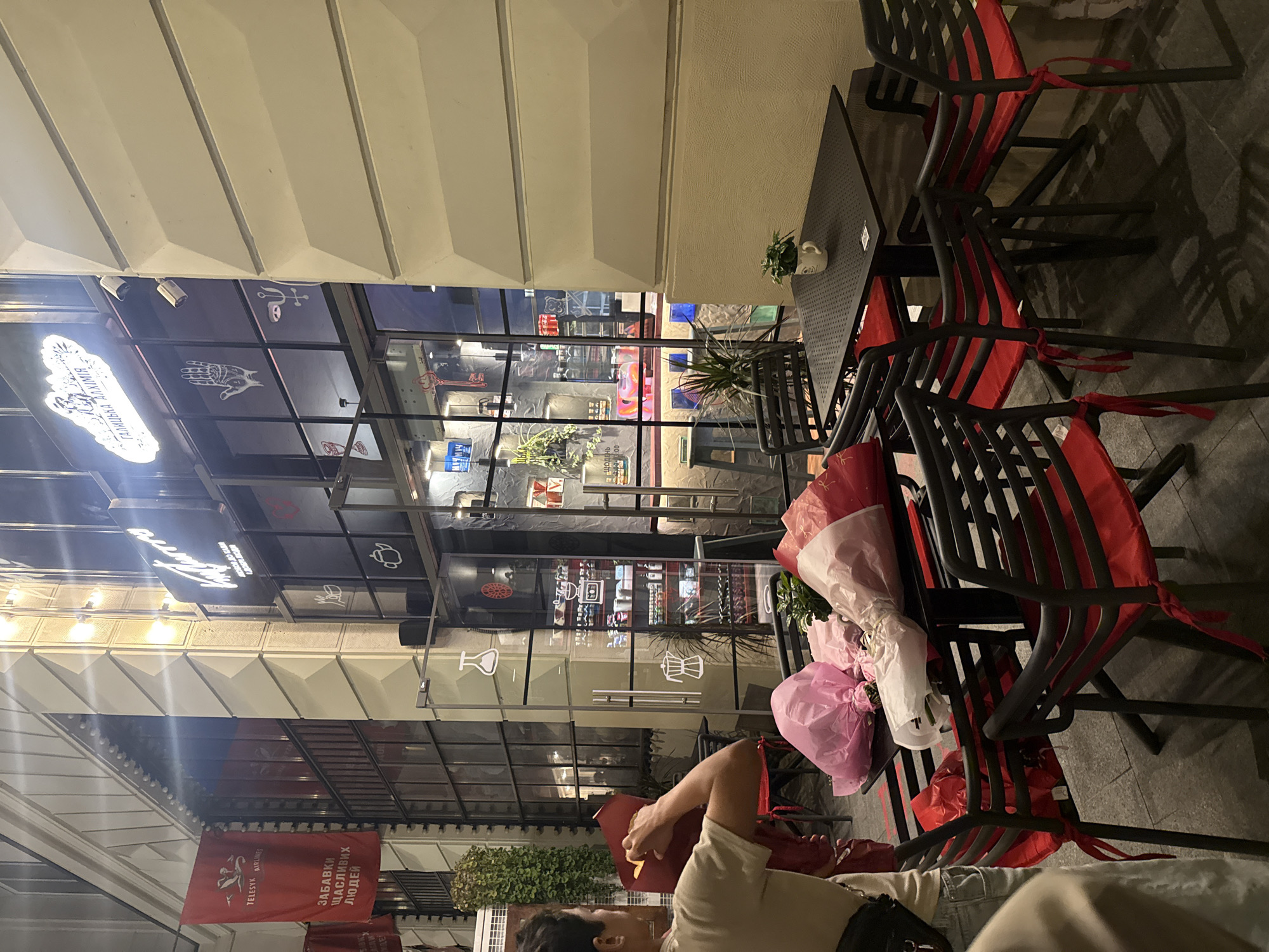 The image captures an outdoor seating area at night, likely in Lviv, Ukraine, given the signage. The focus is on several tables and chairs with red cushions, some adorned with bouquets wrapped in pink and white paper. A woman is visible on the left side of the frame, in the process of handling a bouquet. Behind the seating area is the facade of a building with large windows, revealing the interior of a establishment. Neon signs with Cyrillic lettering are prominently displayed on the building. The lighting suggests it is evening, with artificial lights illuminating the scene.