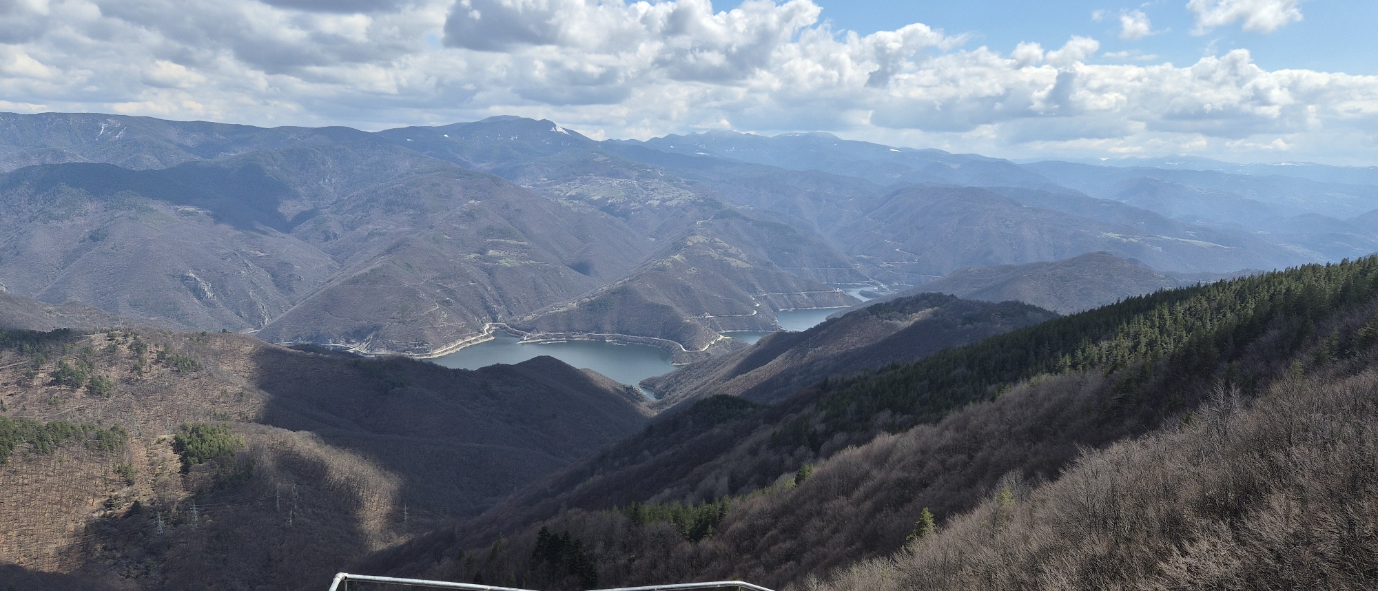 The image shows a mountainous landscape in Ravnogor, Bulgaria, under a partly cloudy sky. The foreground is dominated by hills with sparse vegetation, transitioning to denser coniferous forests on some slopes. In the midground, a large, winding lake stretches through the valleys between the mountains. In the background, several mountain ranges fade into the distance, with some snow visible on the higher peaks. The light casts shadows across the terrain, creating contrast and emphasizing the topography.