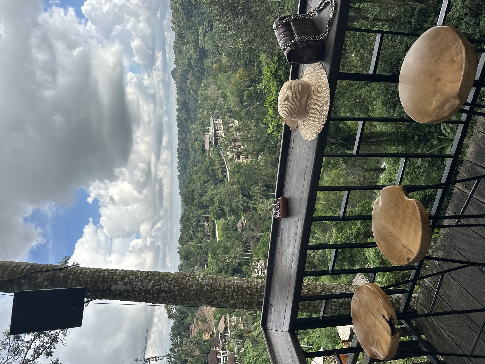 A view from a high vantage point overlooking a lush, green landscape in the Ubud District, Indonesia. The scene is captured from a balcony or elevated structure with a wooden railing.
Several wooden stools with organic, irregular shapes are attached to the railing. On the railing itself, a straw hat and a dark-colored handbag with a chain strap rest. A small, rectangular brown object is also positioned on the railing.
In the background, a dense forest covers rolling hills, interspersed with residential buildings and villas. The sky is partly cloudy, with patches of blue visible amidst the overcast conditions. A tree trunk, possibly part of the structure, is visible with a rectangular black object attached.