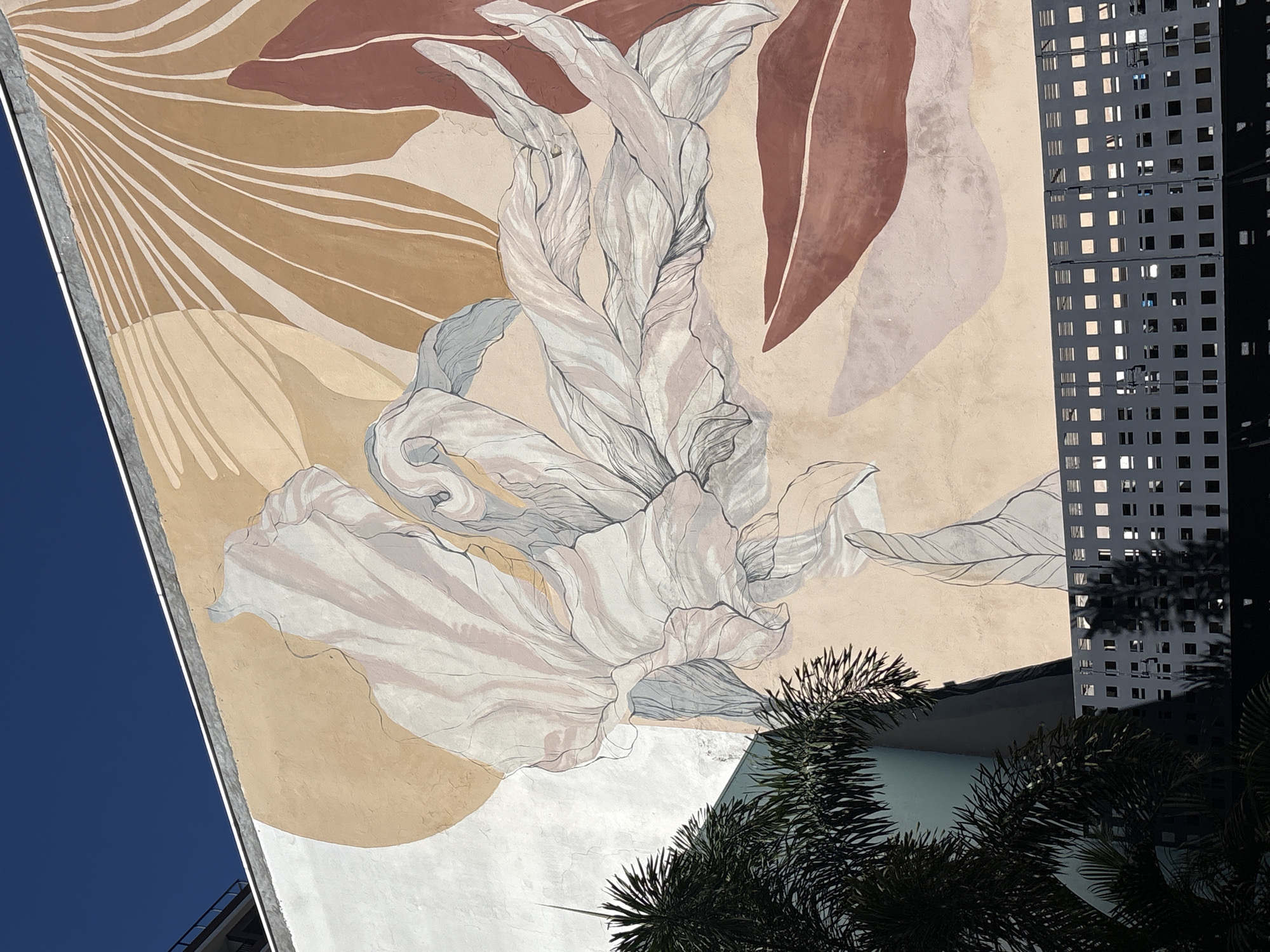 A wide-angle, upward-looking shot captures a building with a large mural painted on its side, set against a clear, deep blue sky. The mural features abstract floral or leaf-like shapes in soft, earthy tones of beige, pale pink, and rust-red, outlined with fine black lines. To the left of the mural, a dark green palm tree with spiky fronds partially obscures the building's facade, which is a muted blue-green. In the foreground, a dark grey metal fence with a repeating square-and-line pattern runs horizontally across the bottom of the frame, casting shadows from unseen elements. A portion of another building, dark and topped with railings, is visible at the top left corner, suggesting an urban setting. The mural's style is reminiscent of art found in the Vila Madalena neighborhood of São Paulo, Brazil, known for its vibrant street art and bohemian atmosphere. No people are visible in the image.