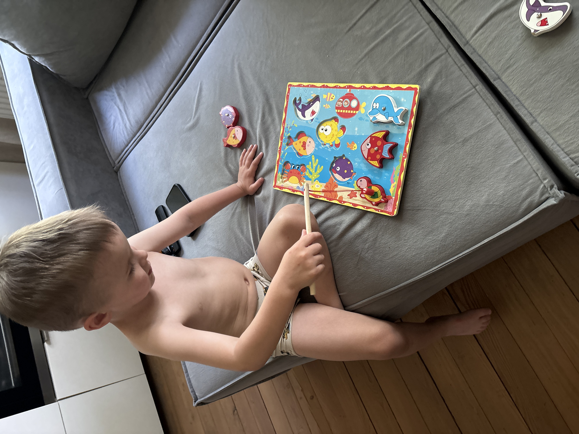 A young, shirtless boy is sitting on a gray couch, playing with a wooden puzzle featuring sea creatures. The puzzle is positioned on the couch next to him, with several puzzle pieces removed and placed near his hand. He is holding a wooden stick. A mobile phone and a TV remote are also on the couch next to him. The couch is situated on a wooden floor.
There is nothing in the picture to place the location to the city of Chaikovsky, Russia.