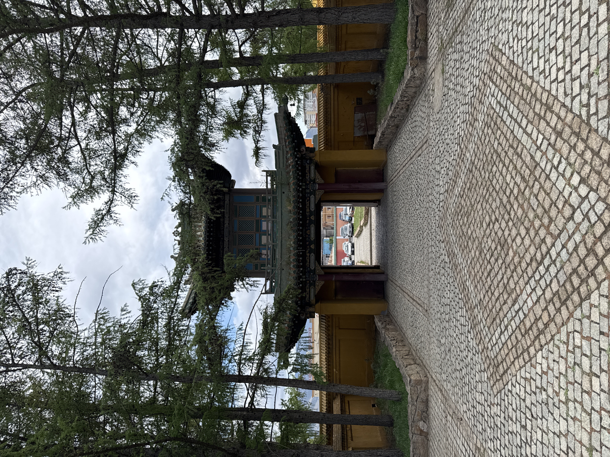 The image depicts the entrance to the Choijin Lama Temple in Ulaanbaatar, Mongolia, viewed from a cobblestone path. A traditional gate with a multi-tiered roof, decorated with intricate patterns and colors, stands between tall trees. The walls surrounding the temple are yellow, and there is a glimpse of modern buildings and vehicles through the gate. The sky is cloudy.