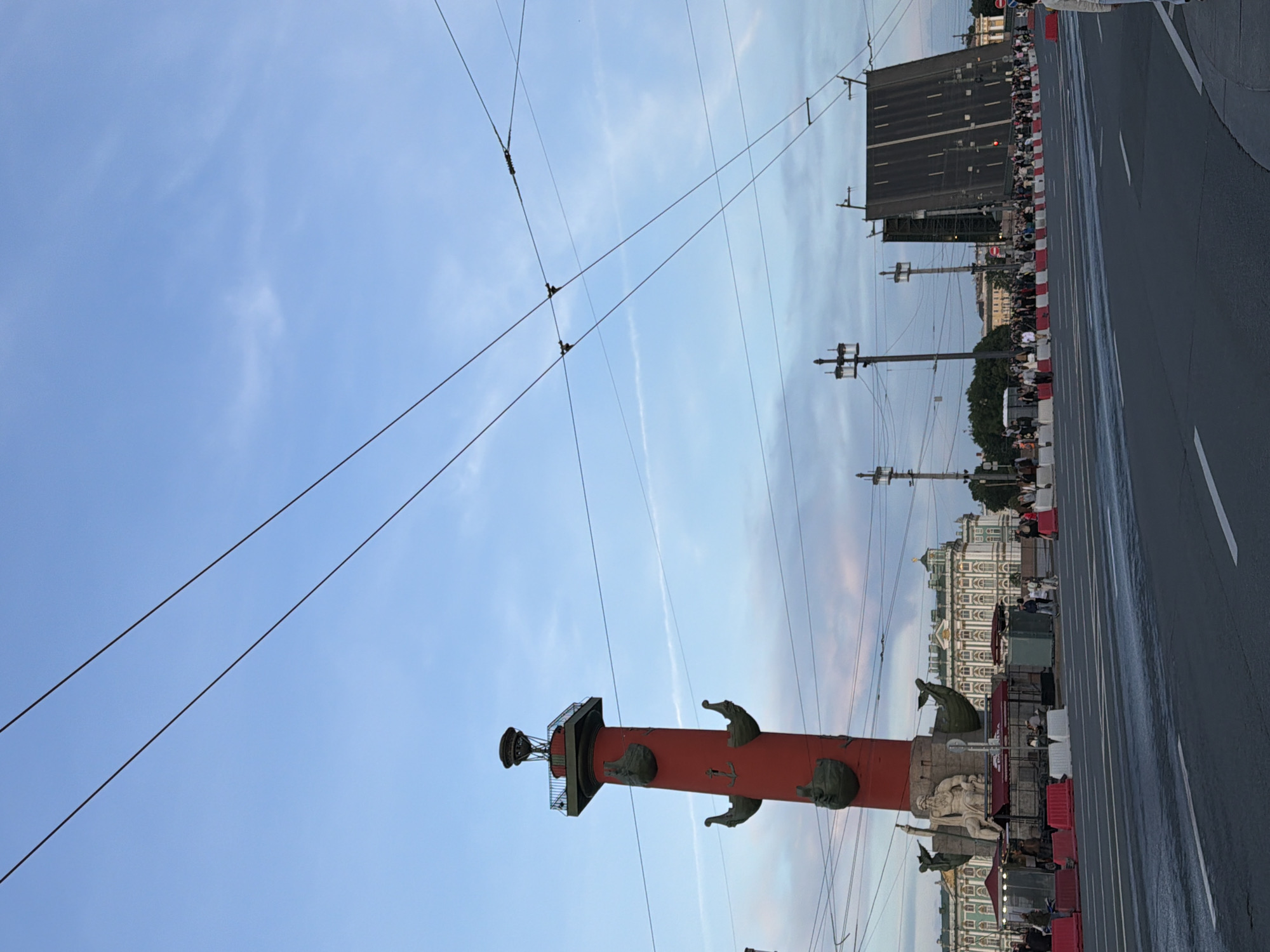 This is a medium-range, eye-level, outdoor shot of the Rostral Column in Saint Petersburg, Russia. The column is red and topped with a lantern. It is decorated with three anchor-shaped reliefs. At the base of the column is a statue of a seated man, possibly Neptune, holding a trident. In the background, a large, ornate building, possibly the State Hermitage Museum, is visible. To the right, a large drawbridge is raised, with a crowd of people gathered on the embankment. The sky is blue with some wispy clouds, and several overhead wires are visible. The street in the foreground is asphalt with white lane markings.