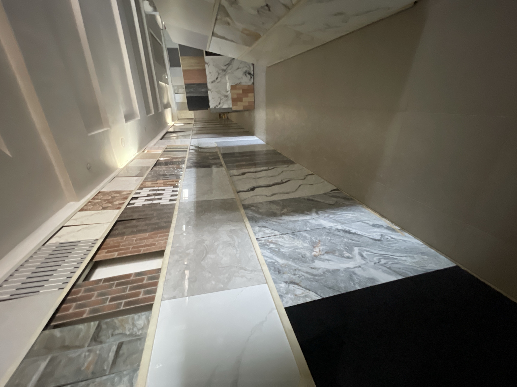 The image depicts a display of various tile samples in what appears to be a showroom or warehouse setting. The tiles are arranged in rows, showcasing different patterns, colors, and materials, including stone, brick, and ceramic. The lighting is bright and the perspective is distorted, giving a narrow view. The location is likely in Abuja, Nigeria, given the user's prompt, but there is no specific evidence in the image to confirm the exact location or identify any individuals.