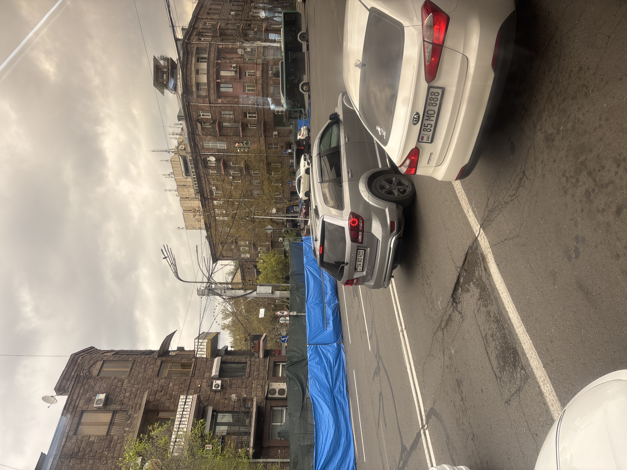 The image shows a street scene in Yerevan, Armenia. There are several cars parked on the side of the road, including a white Kia Forte and a silver SUV with license plate "14 AO 411." A bus is parked further up the street. A blue tarp is stretched along the side of the road, likely indicating ongoing construction or maintenance work. The buildings on either side of the street are older, multi-story structures with a mix of residential and commercial spaces. The sky is overcast. The photo appears to have been taken from inside a vehicle.