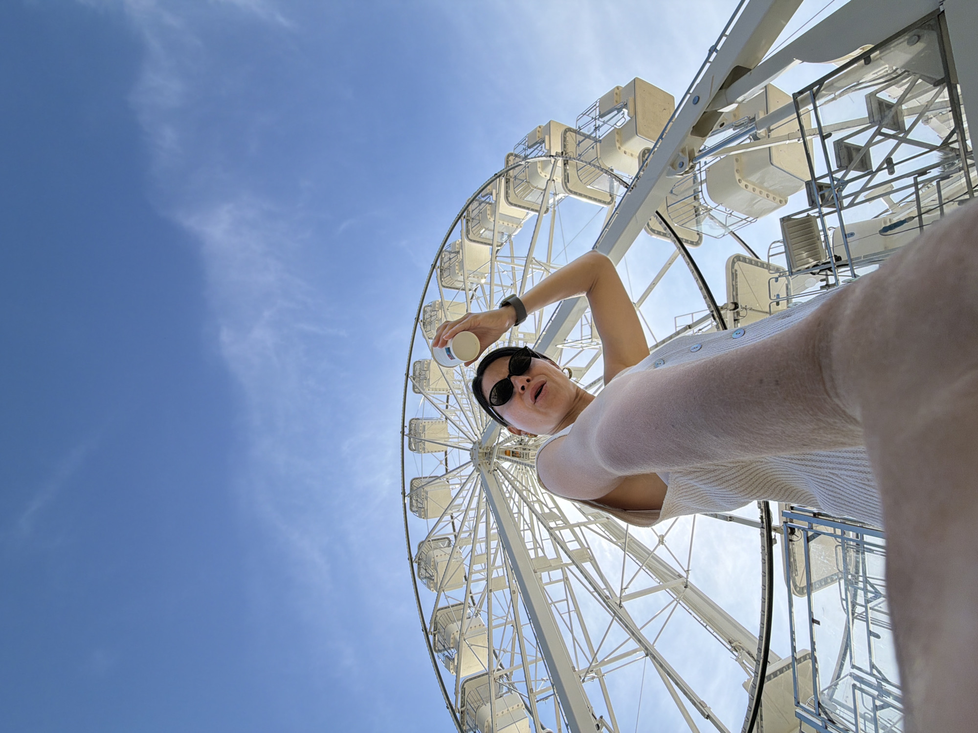 A low-angle shot shows a woman holding a small white container, possibly food or drink, against a bright blue sky partially obscured by a large, white Ferris wheel. The woman is wearing sunglasses and a watch, and her arm is raised, positioning the container above her. The Ferris wheel's structure is white, and its enclosed cabins are visible. The sky is clear except for faint wisps of clouds. The scene appears to be in Budva, Montenegro, given the likely presence of the Ferris wheel in that city.