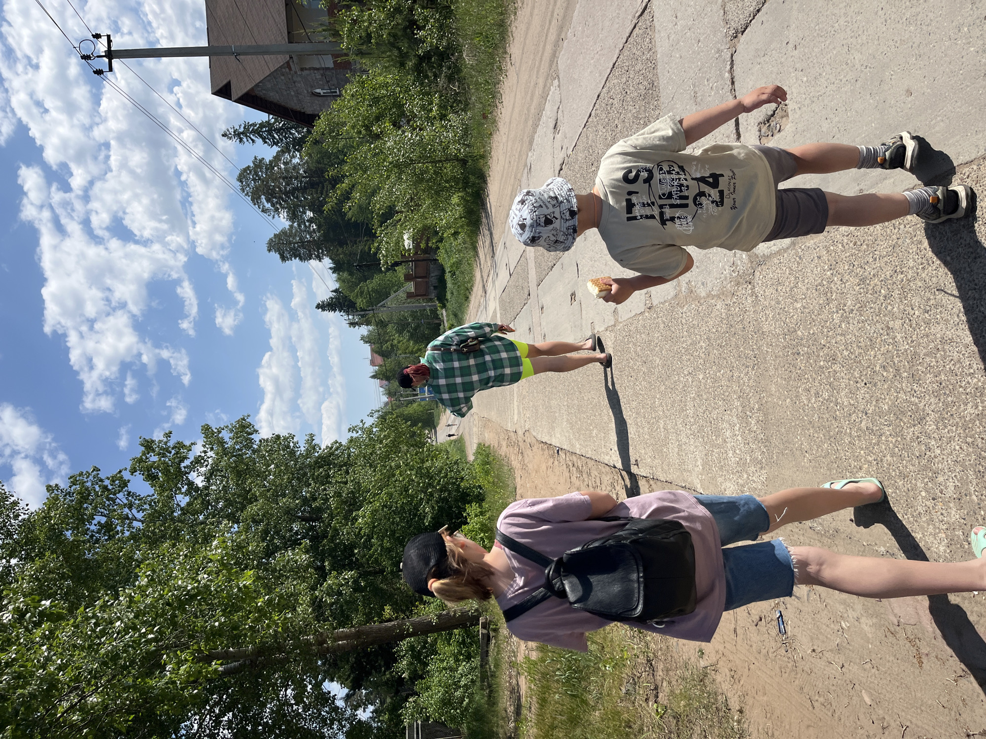 Three individuals are walking on a concrete sidewalk in Chaikovsky, Russia. The individual closest to the viewer wears a pink t-shirt, denim shorts, a black baseball cap, and a black backpack. The second individual wears a green and white checkered shirt and bright yellow shorts. The third individual, furthest away, wears a white bucket hat, a light-colored t-shirt, and grey shorts; they hold something in their hand. The background features trees, a blue sky with scattered clouds, and a building with a power pole. The ground is a mix of concrete and dirt with sparse vegetation.
