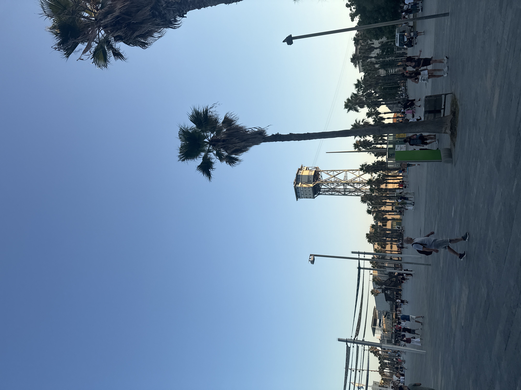 A wide-angle, low-angle shot captures a vibrant public space in Barcelona, Spain, likely near the Port Vell area, under a clear blue sky. The foreground is dominated by a grey paved area with a man in a grey t-shirt and shorts walking towards the right. Palm trees, with their distinctive fronds, are scattered throughout the scene, with one particularly tall specimen in the mid-ground.
In the background, a distinctive industrial tower, possibly a historic structure related to the port, stands tall. Beyond it, a row of modern buildings and more palm trees line the landscape. Several people are seen strolling in the plaza, with groups of individuals and smaller clusters of people visible. Some are walking casually, while others appear to be interacting in small groups. Streetlights, some with distinctive orange accents, are positioned throughout the plaza, indicating it's a well-lit public area. The scene suggests a pleasant, active atmosphere, typical of a tourist-friendly urban environment.