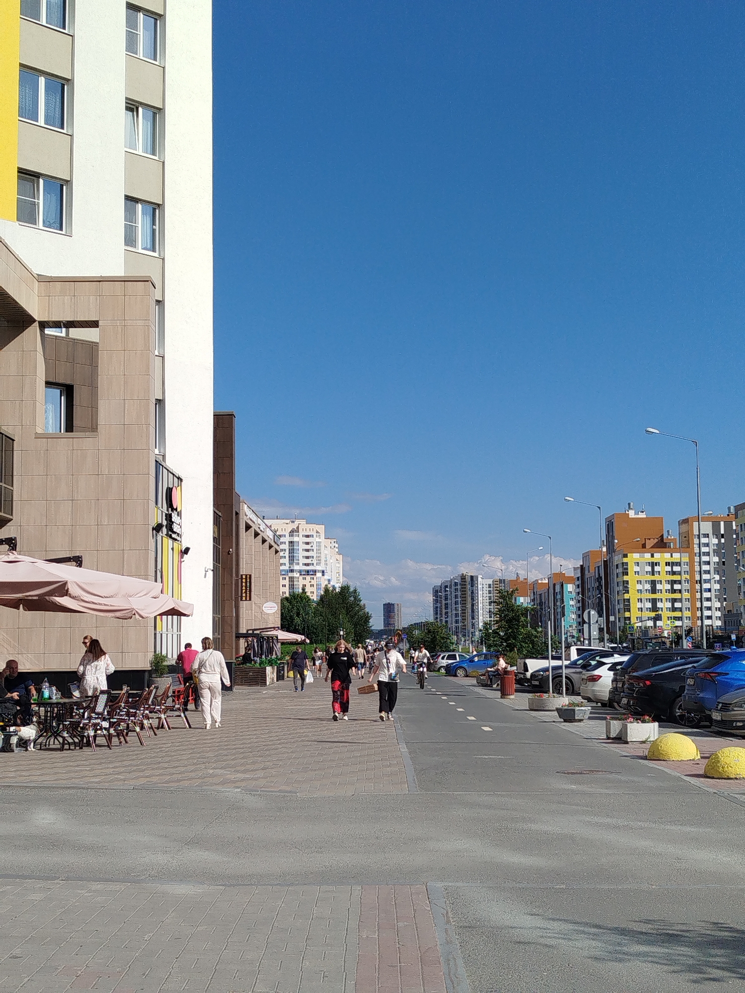 The image depicts a street scene in Yekaterinburg, Russia, likely in a newly developed residential area. On the left, a modern multi-story building with a light-colored facade and yellow accents dominates the foreground. An outdoor seating area with tables and chairs, some covered by beige umbrellas, is situated in front of the building. People are seen walking along the sidewalk and sitting at tables. The street is lined with parked cars on the right side. Further down the street, a row of colorful apartment buildings of varying heights can be observed, characteristic of contemporary urban development. The sky is clear and blue.