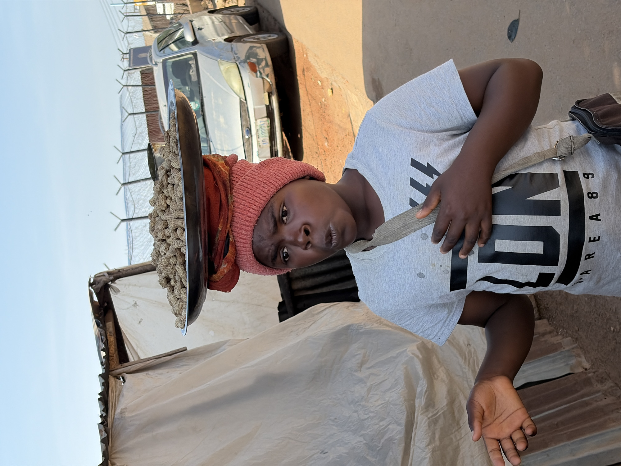 A young Black person wearing a pink knitted beanie and a light gray t-shirt with a large black graphic is standing outdoors in what appears to be a market area in Gura Top, Nigeria. They are balancing a large metal tray filled with peanuts on their head, with a red fabric bundle underneath the tray. The person is looking towards the viewer with an open mouth, as if speaking or surprised. Their left arm is extended outwards, palm open, while their right hand rests on their chest. A light brown strap goes across their chest, supporting a dark bag at their side. In the background, there is a makeshift stall with a corrugated metal roof and light-colored sheeting. Behind the stall and to the right, a silver car is parked on a dirt road, with a fence and some wires visible further in the distance. The overall impression is of a street vendor in a bustling urban environment.