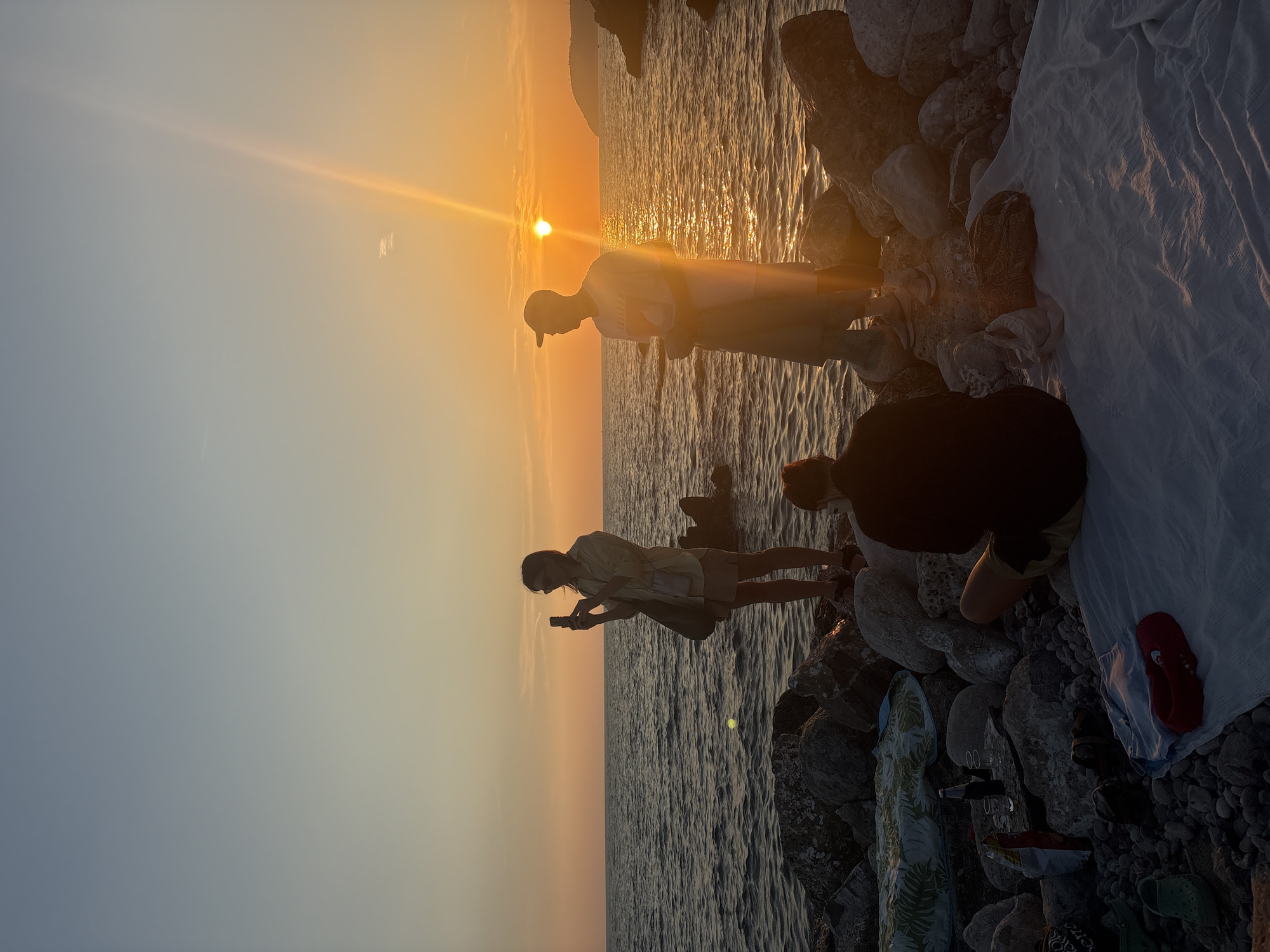 A group of people are gathered on a rocky shore overlooking the sea at sunset. The sky is a gradient of soft blues and oranges, with the sun casting a warm glow across the water. A woman in the center of the frame is holding up a smartphone, seemingly taking a photograph of the sunset. To her right, a man in a t-shirt and cap is looking out at the sea. In the foreground, a person is sitting on a white blanket, facing away from the camera. Scattered around are a few inflatables, a bottle, and some snacks, suggesting a casual gathering. The rocky terrain and the sea are characteristic of the coastal areas of Bar, Montenegro.